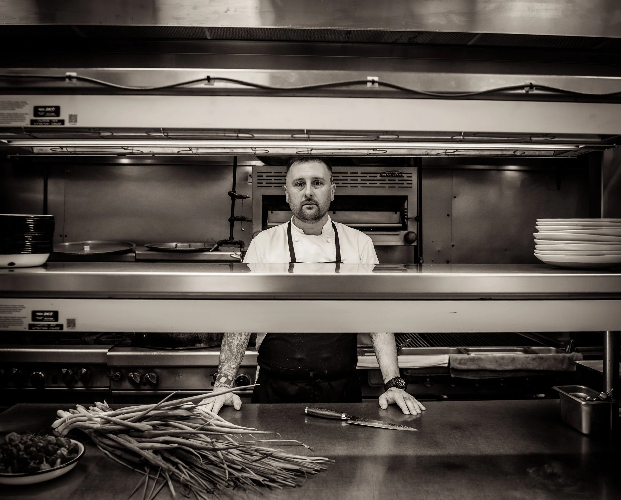 Black-and-white photo of a male chef standing behind a kitchen counter, with a bunch of vegetables and a small bowl of berries in front. The chef is wearing a white apron and has tattoos on his arms, with plates and kitchen equipment in the background.