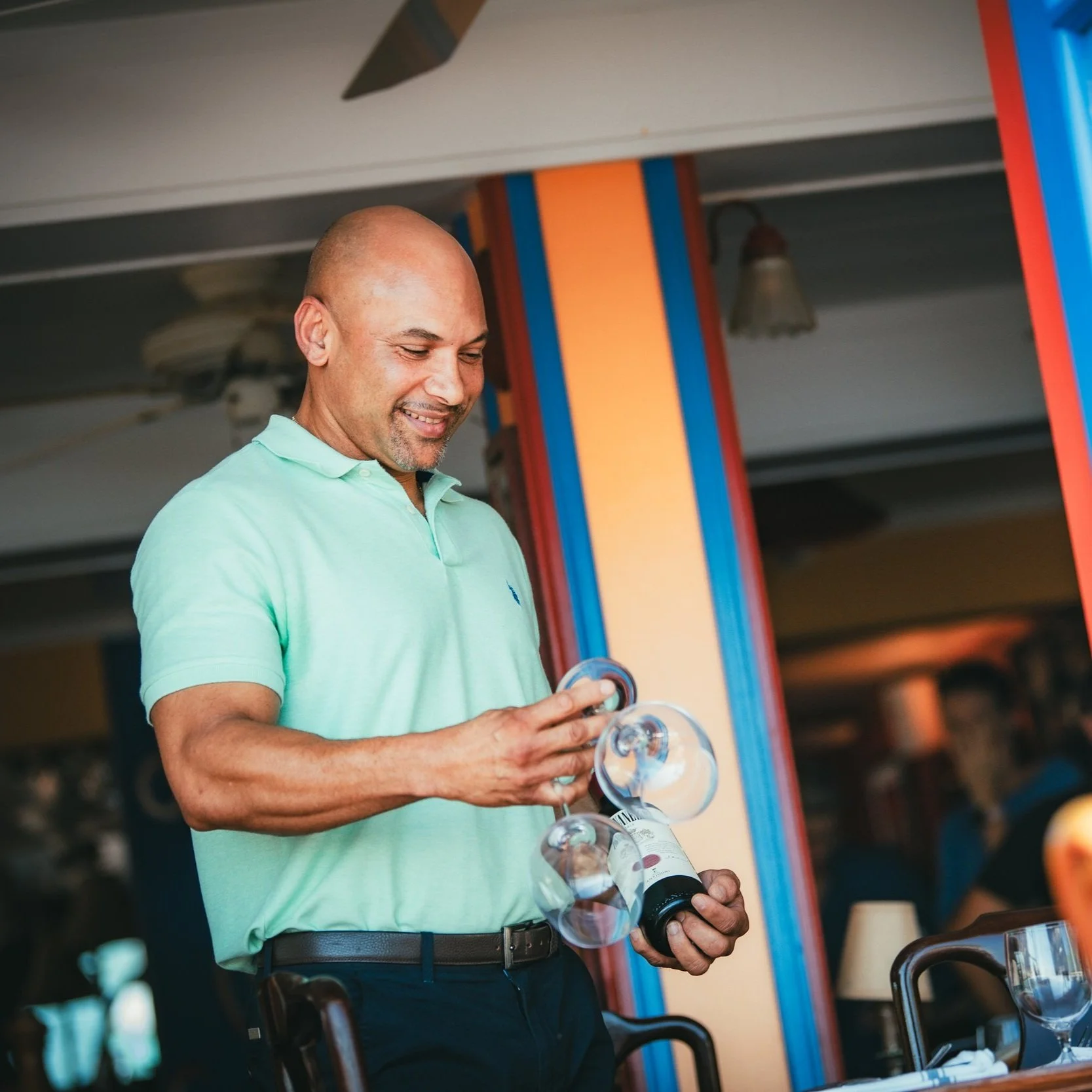 A man wearing a light green polo shirt pours wine into two glasses at a restaurant.