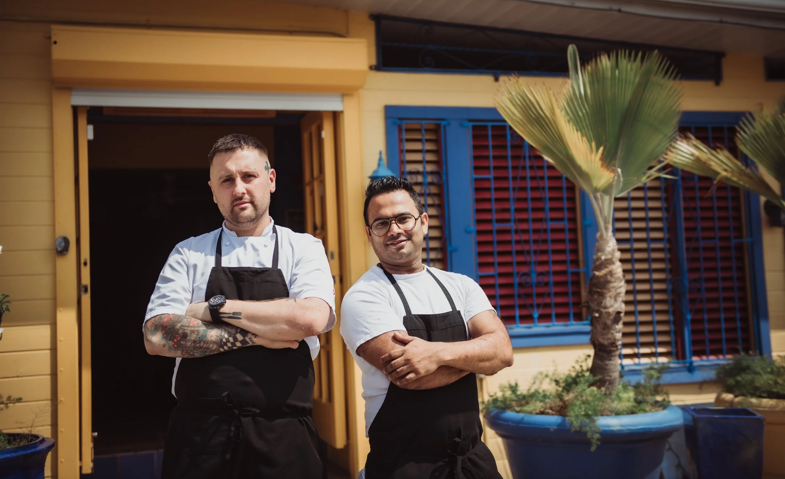 Two male chefs standing outside a restaurant, crossing their arms, in front of a yellow building with blue window shutters and a palm tree in a large blue pot.