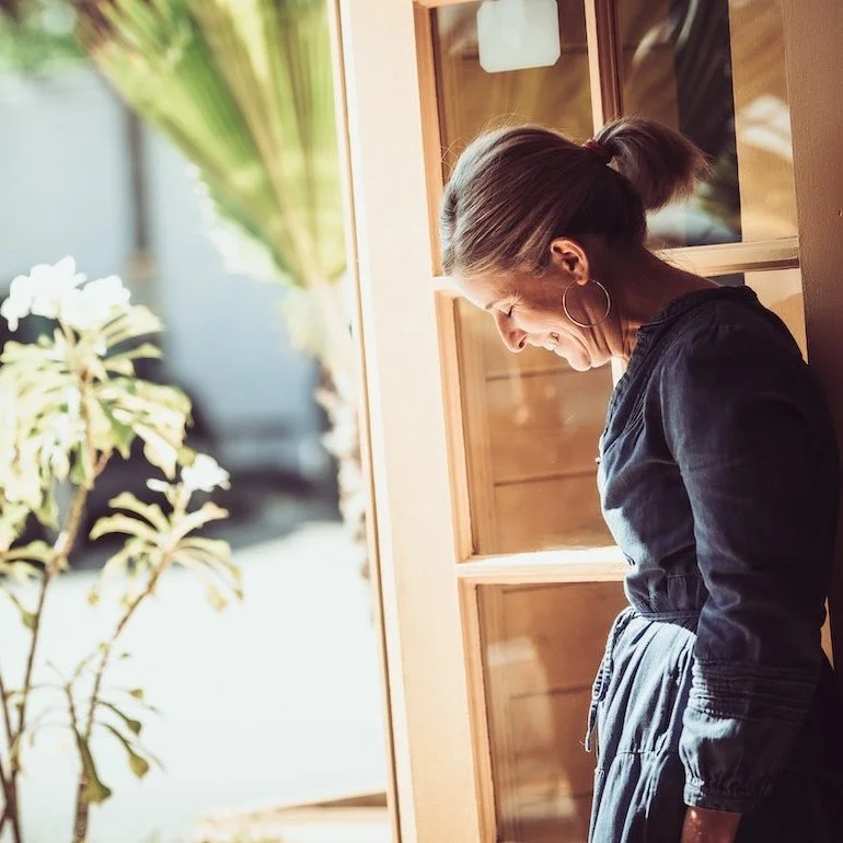 A woman with brown hair in a ponytail, wearing hoop earrings and a dark jumpsuit, stands smiling by a wooden window, looking down in a brightly lit room with plants outside.