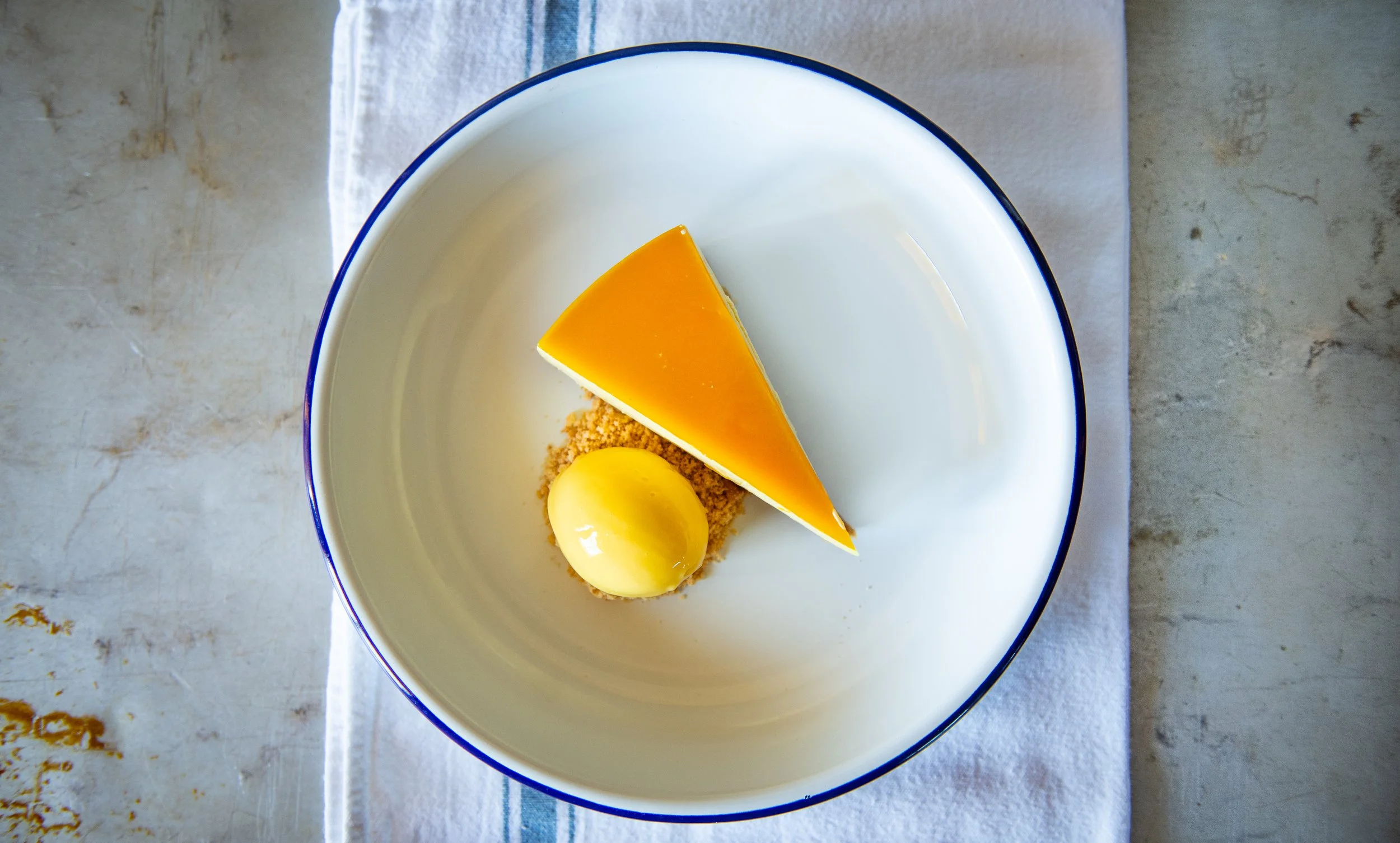 A slice of orange cheesecake on a white plate with a yellow scoop of sorbet or ice cream, on a white cloth napkin.