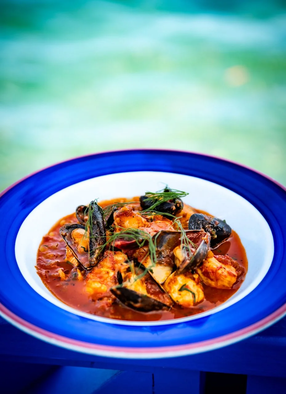 Seafood stew with mussels, fish, and tomatoes in a red broth, garnished with herbs, served in a white bowl with a blue rim.