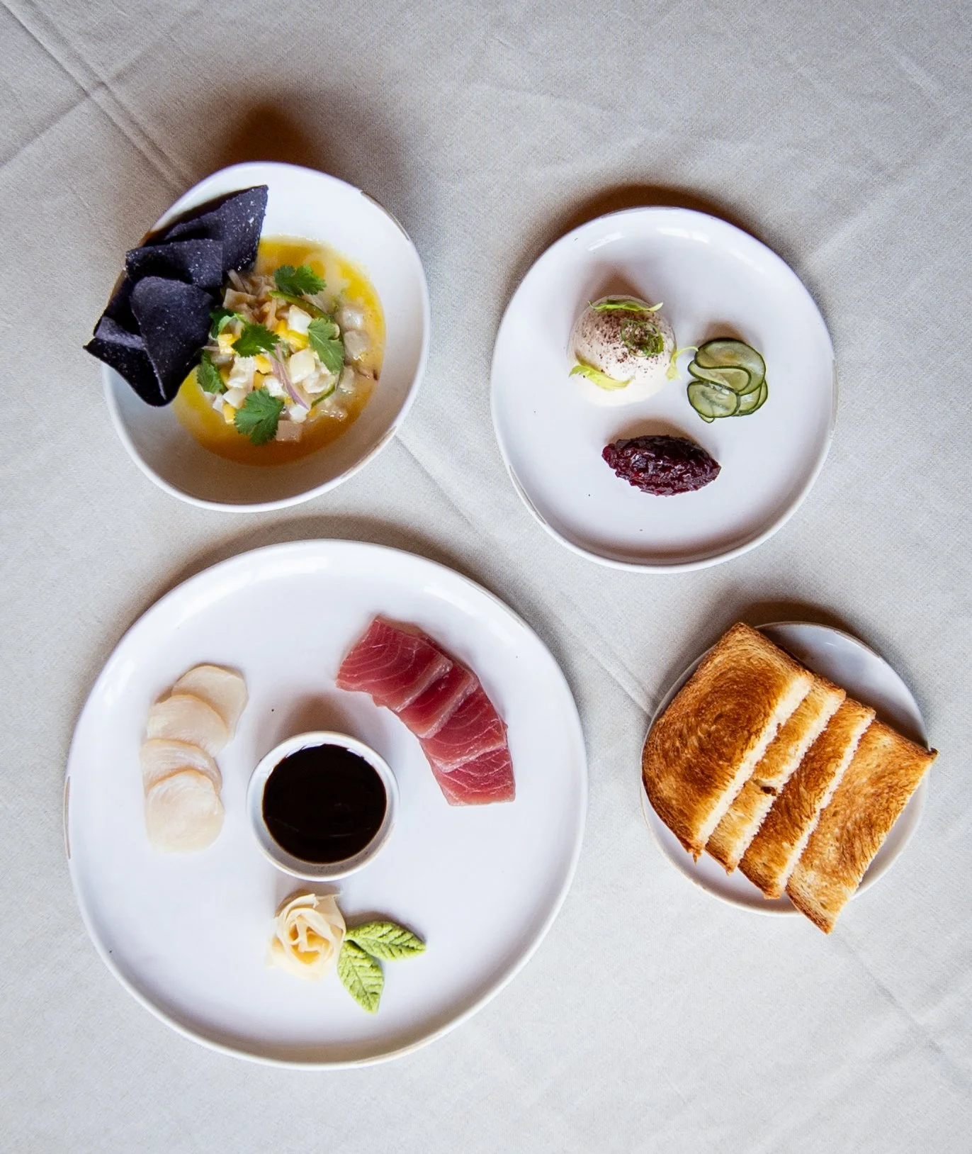 Assorted appetizers and bread on white plates and bowls, including slices of sashimi, dipping sauce, wrapped sushi, and a bowl of salad with chips, and two small dishes with vegetables and a mousse.