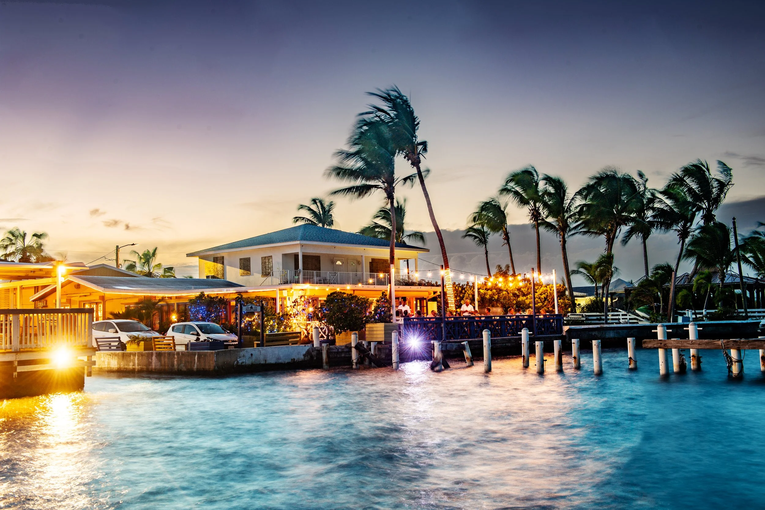 A tropical waterfront scene at dusk with a two-story building, palm trees, cars parked nearby, and string lights illuminating an outdoor patio area.