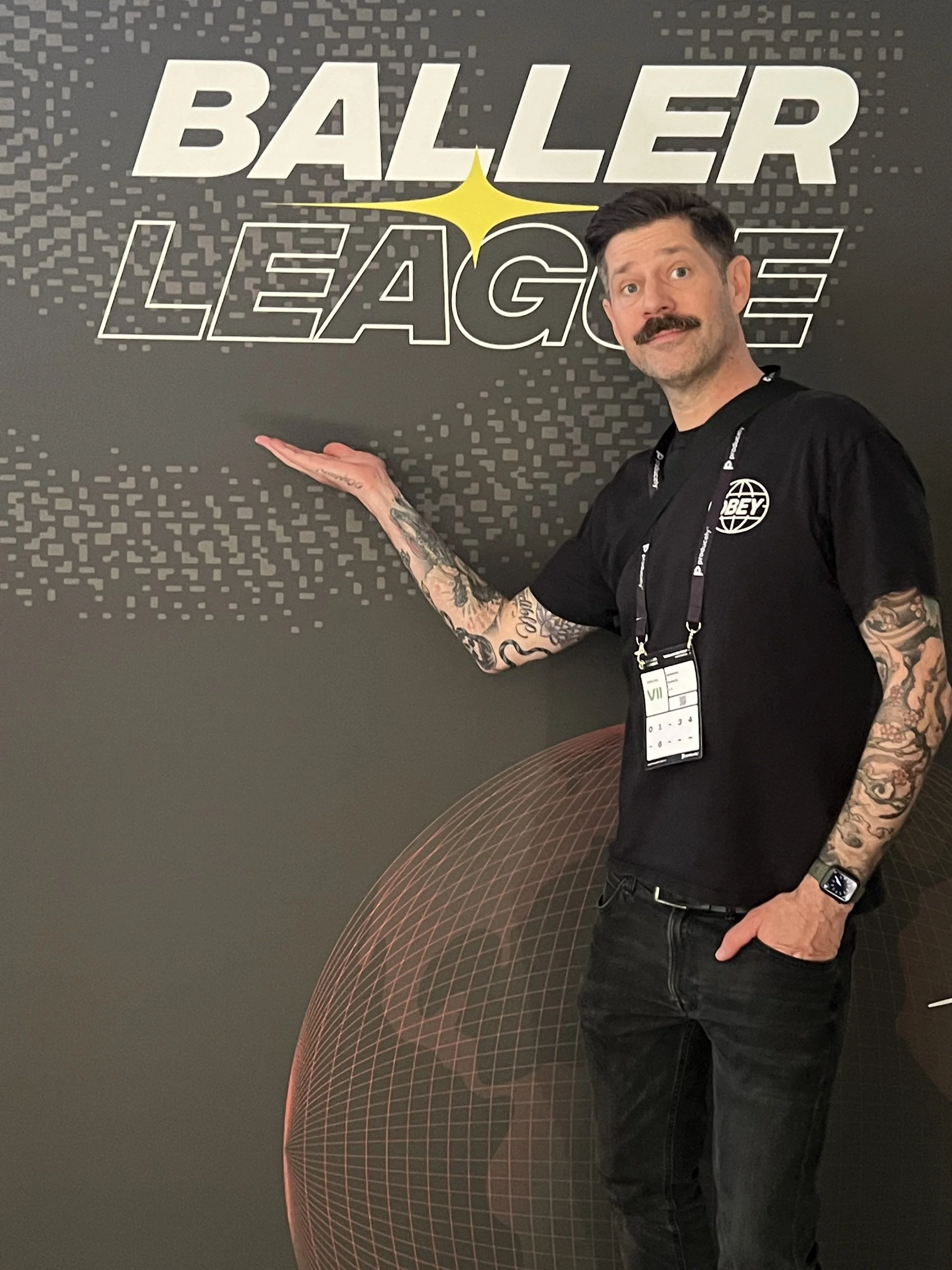 A man with tattoos, mustache, and wearing a black Obey shirt, standing next to a sign that reads 'Baller League' against a black background with digital graphics, pointing to the sign with his left hand.