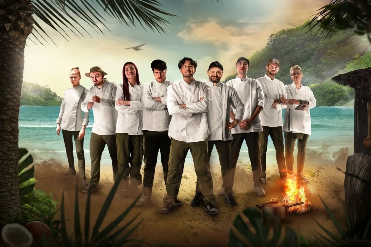 A group of ten chefs in white coats standing on a beach with tropical trees and ocean in the background. There's a small bonfire with a pot on it and an airplane flying in the sky.