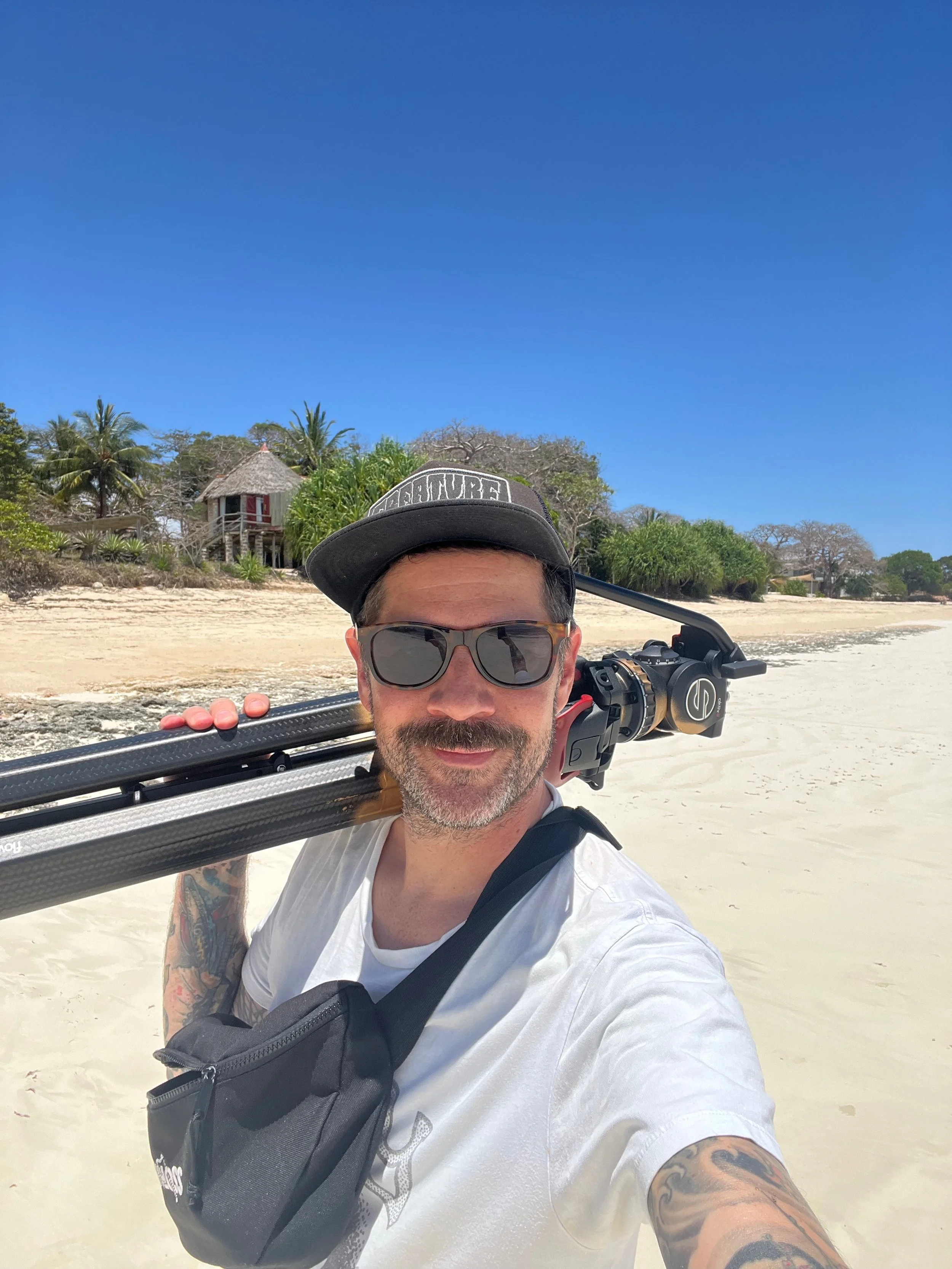 Man with tattoos, sunglasses, and a cap taking a selfie on a beach in Kenya, carrying a tripod over his shoulder, with a background of sandy shoreline, tropical trees, and a thatched-roof hut under a clear blue sky.