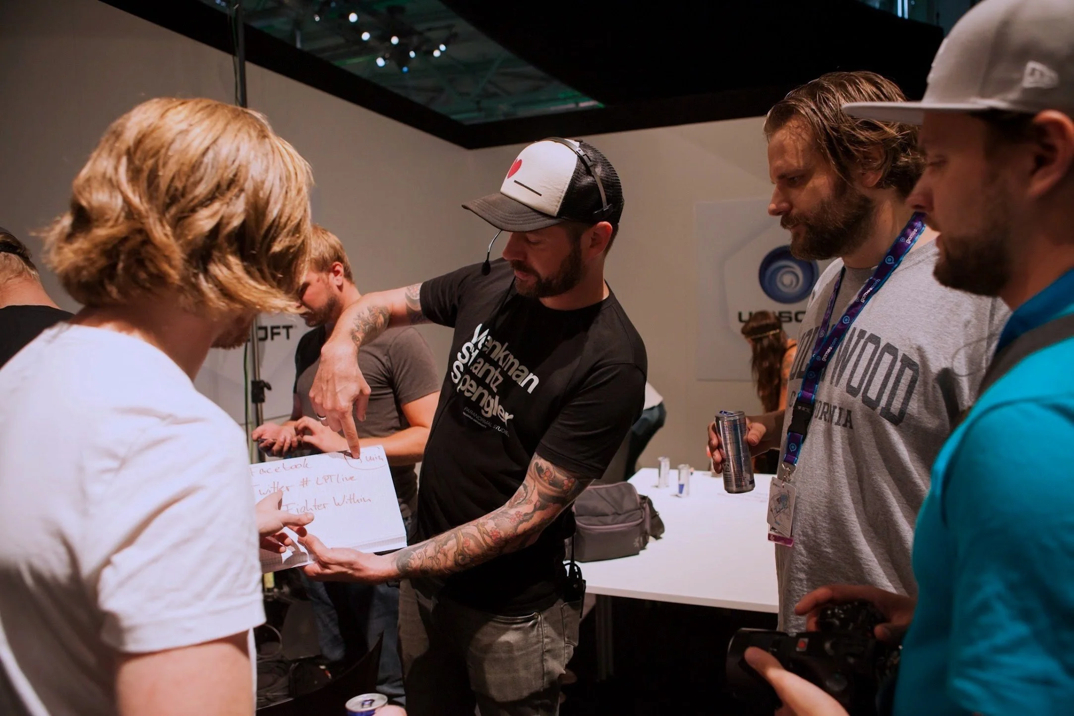 Group of people at a convention or event, with one person showing something on a small whiteboard to others. The background has a sign that reads 'UbiSoft'.