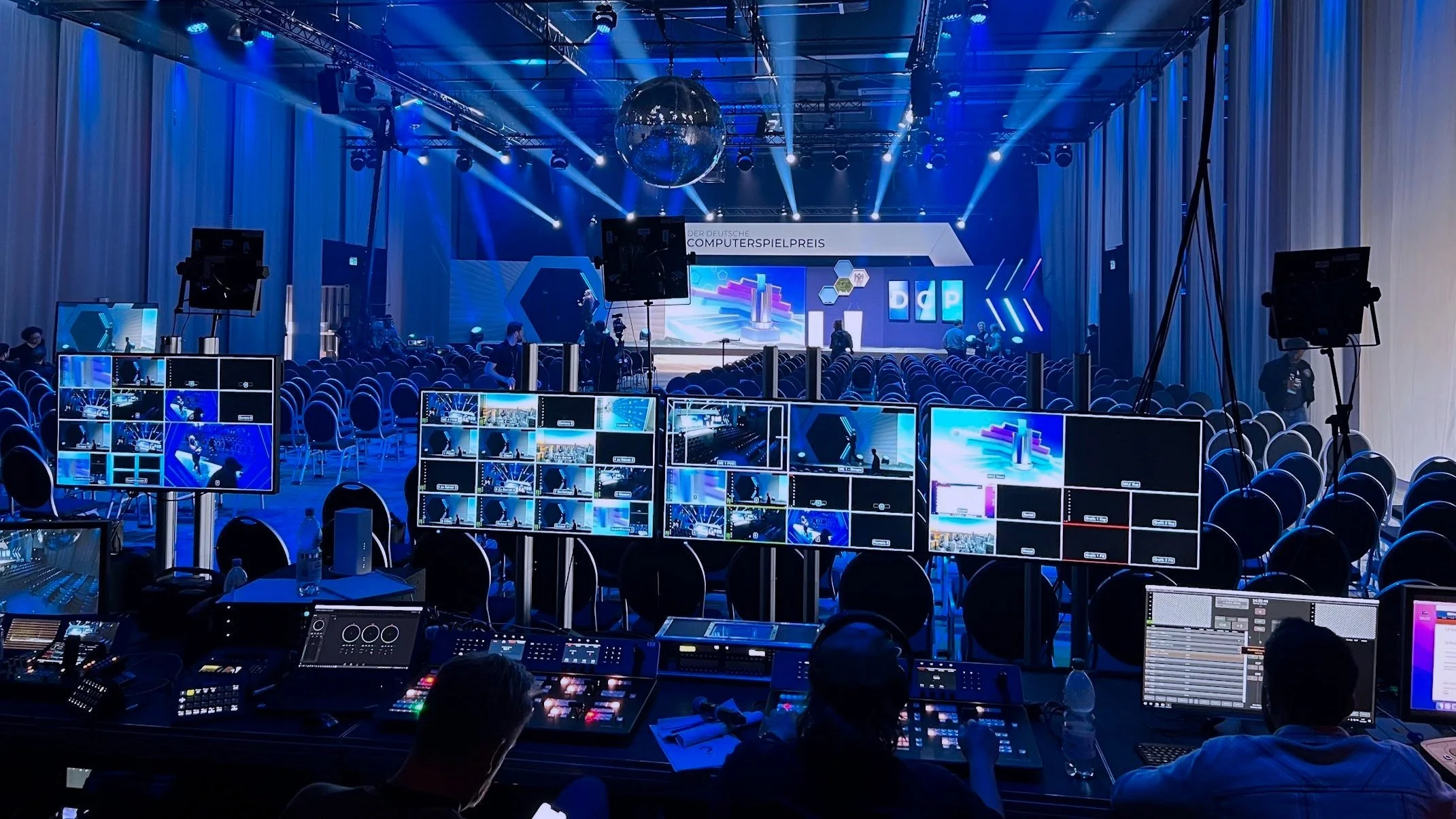 The empty awards ceremony hall of Deutscher Computerspielpreis / DCP with a large stage, multiple monitor screens, professional lighting, and a control desk with audio and video equipment in the foreground.