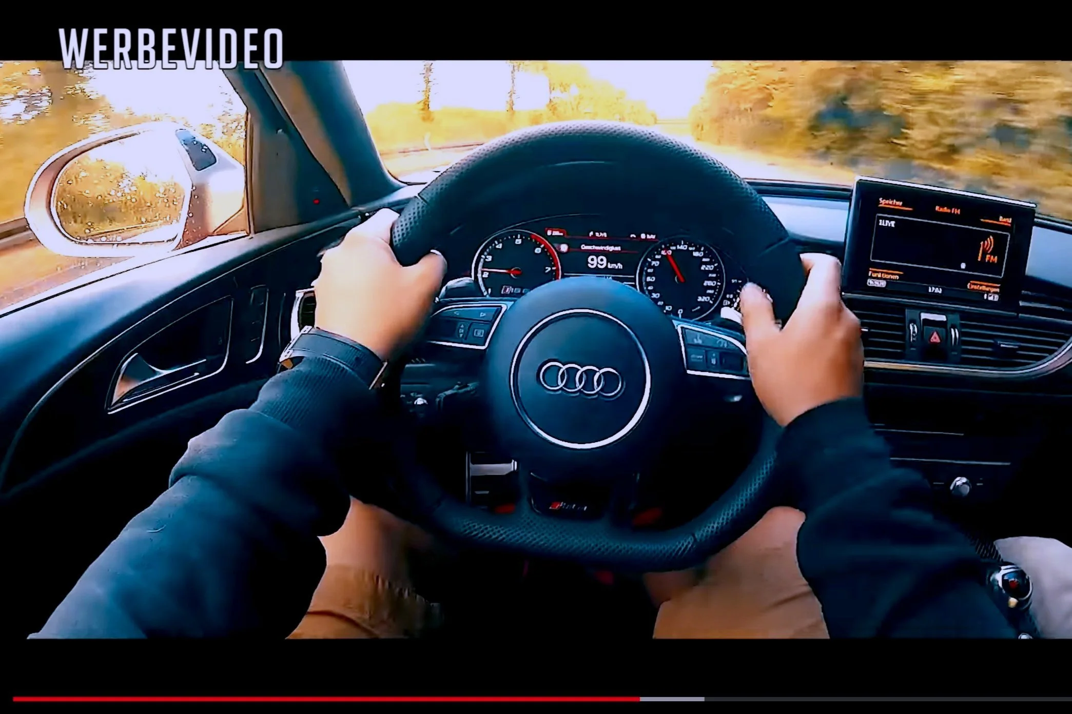 Inside view of an Audi car in motion on a road with a dashboard displaying speed and other controls. The driver is holding the steering wheel with both hands, wearing a watch and a black jacket, with a side mirror reflecting rain on the window.