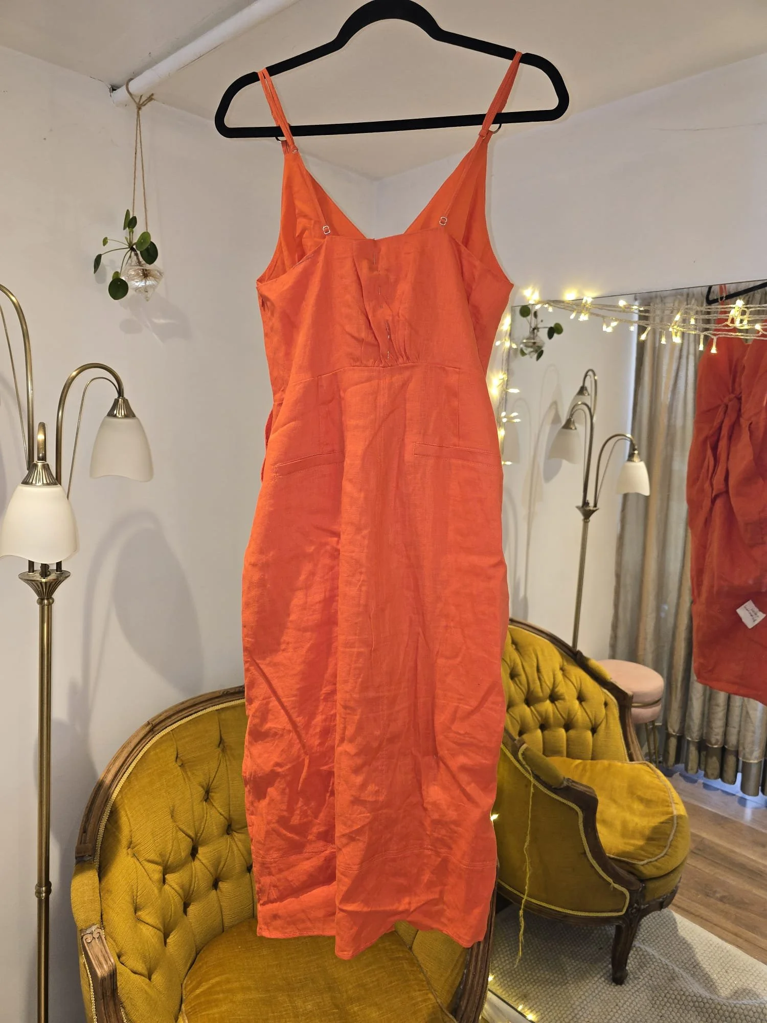 Full-length coral-orange linen sundress photographed before alterations, displayed on a hanger with visible creasing and shaping seams in a Brighton tailoring studio.