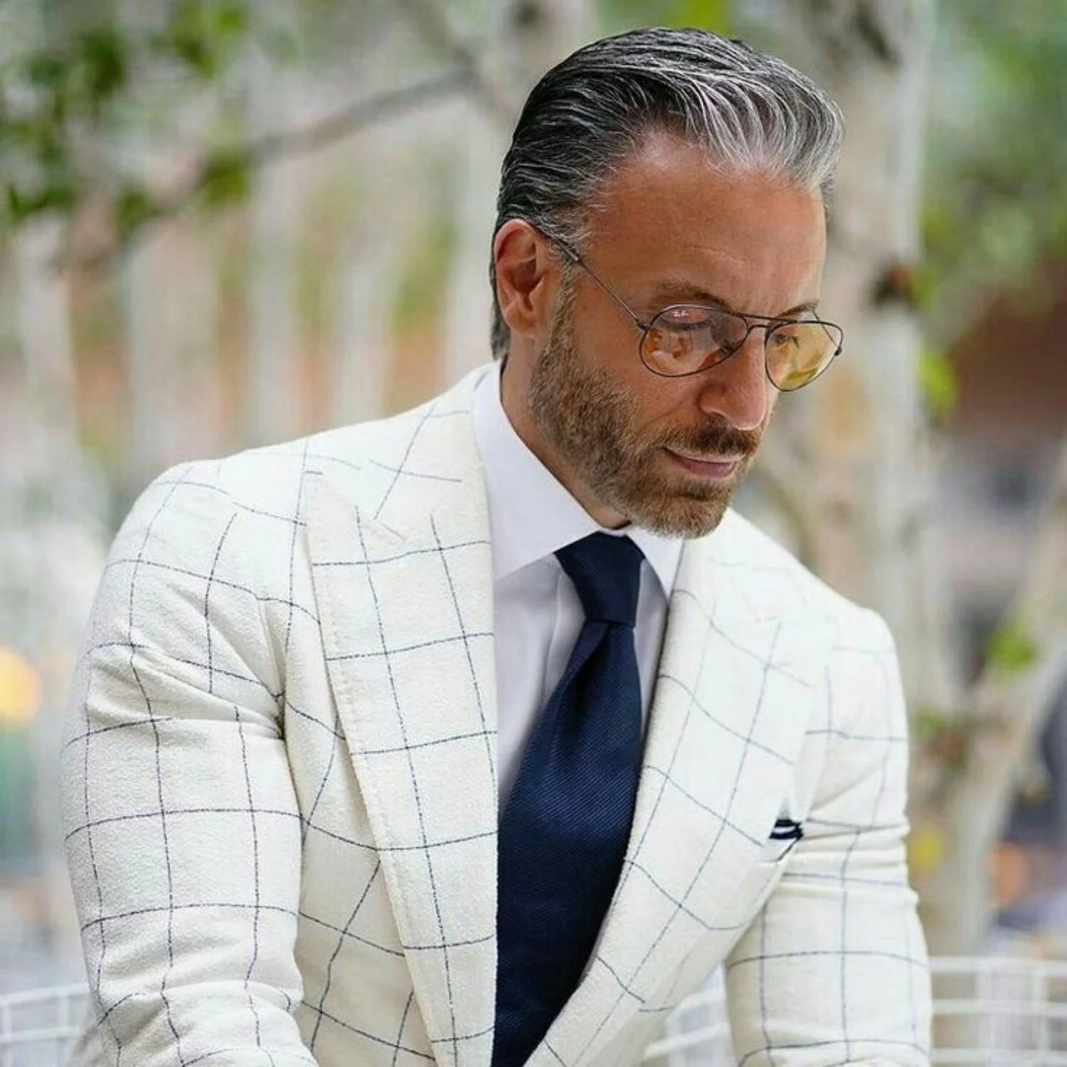 A stylish man with salt-and-pepper hair, beard, and glasses, wearing a white suit jacket with a checkered pattern, white shirt, and dark tie, looking down outdoors with blurred greenery in the background.