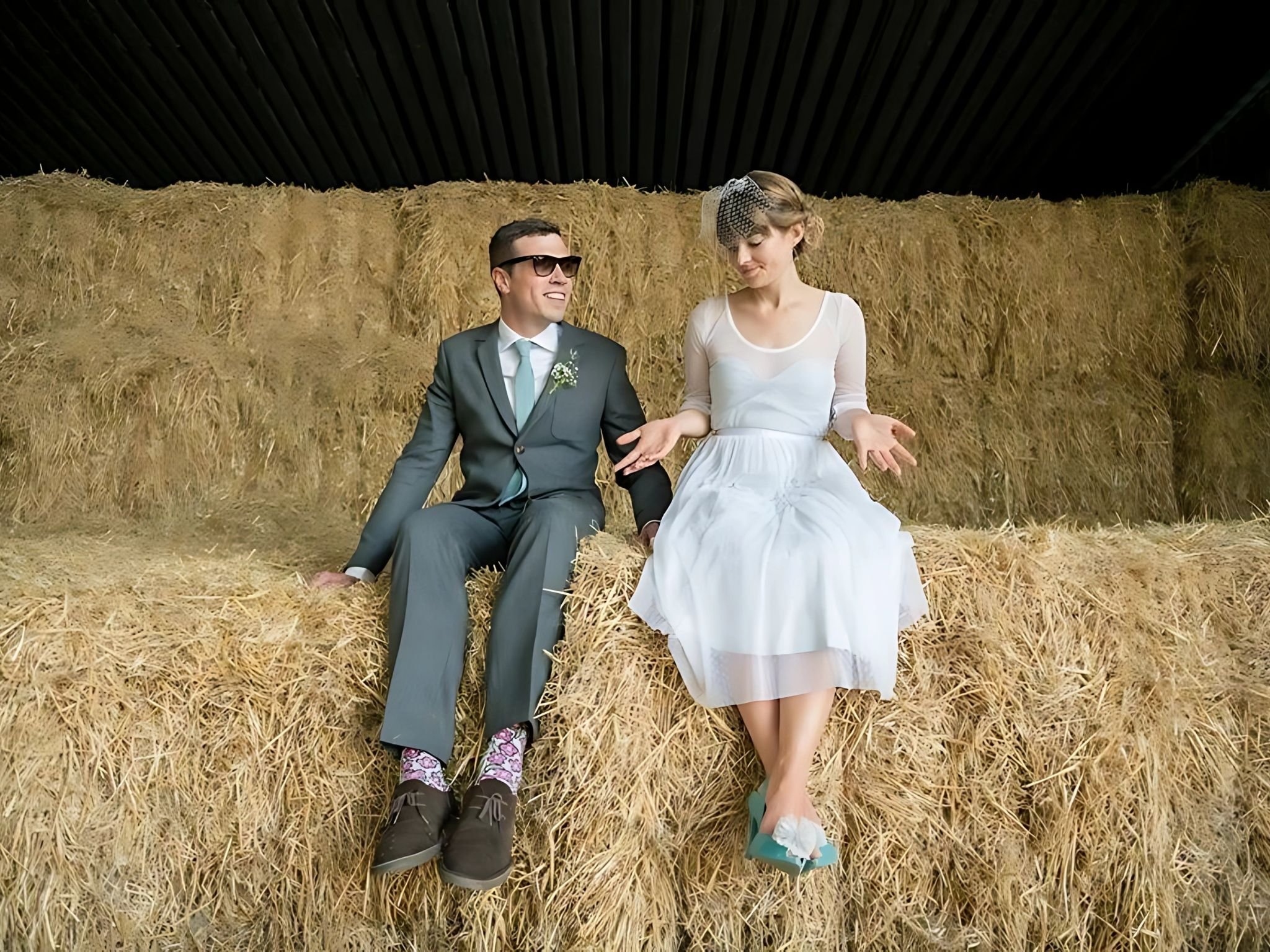 A couple sitting on hay bales, the man in a suit and sunglasses, the woman in a white dress with a birdcage veil, in a barn-like setting.