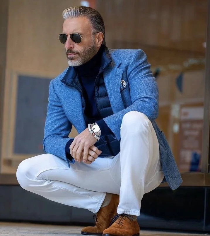 A man crouching, wearing sunglasses, a blue blazer, dark quilted vest, black turtleneck, white pants, and brown boots, with a silver watch and bracelet.