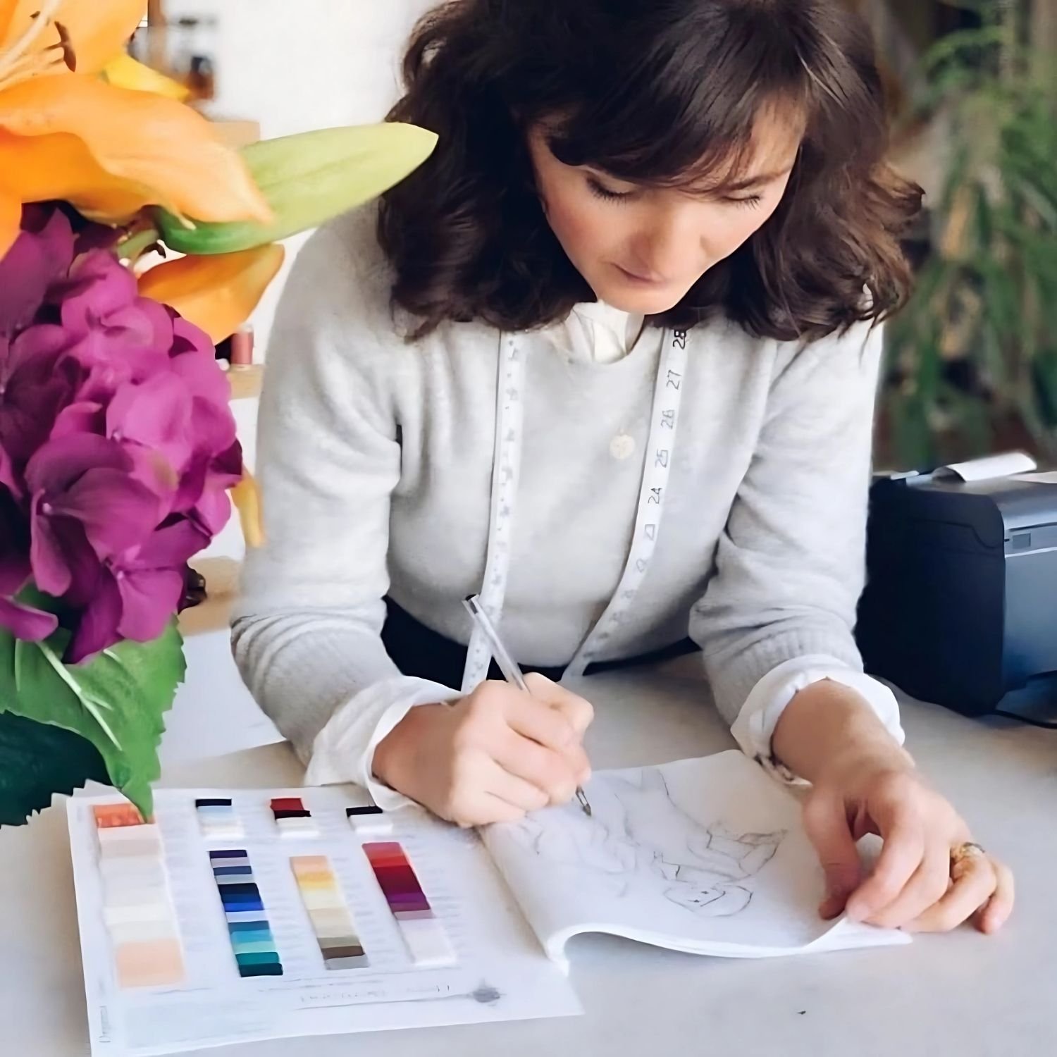 Fashion designer sketching clothes next to a fabric color swatch book, with a tape measure around their neck and a bouquet of flowers on the table.