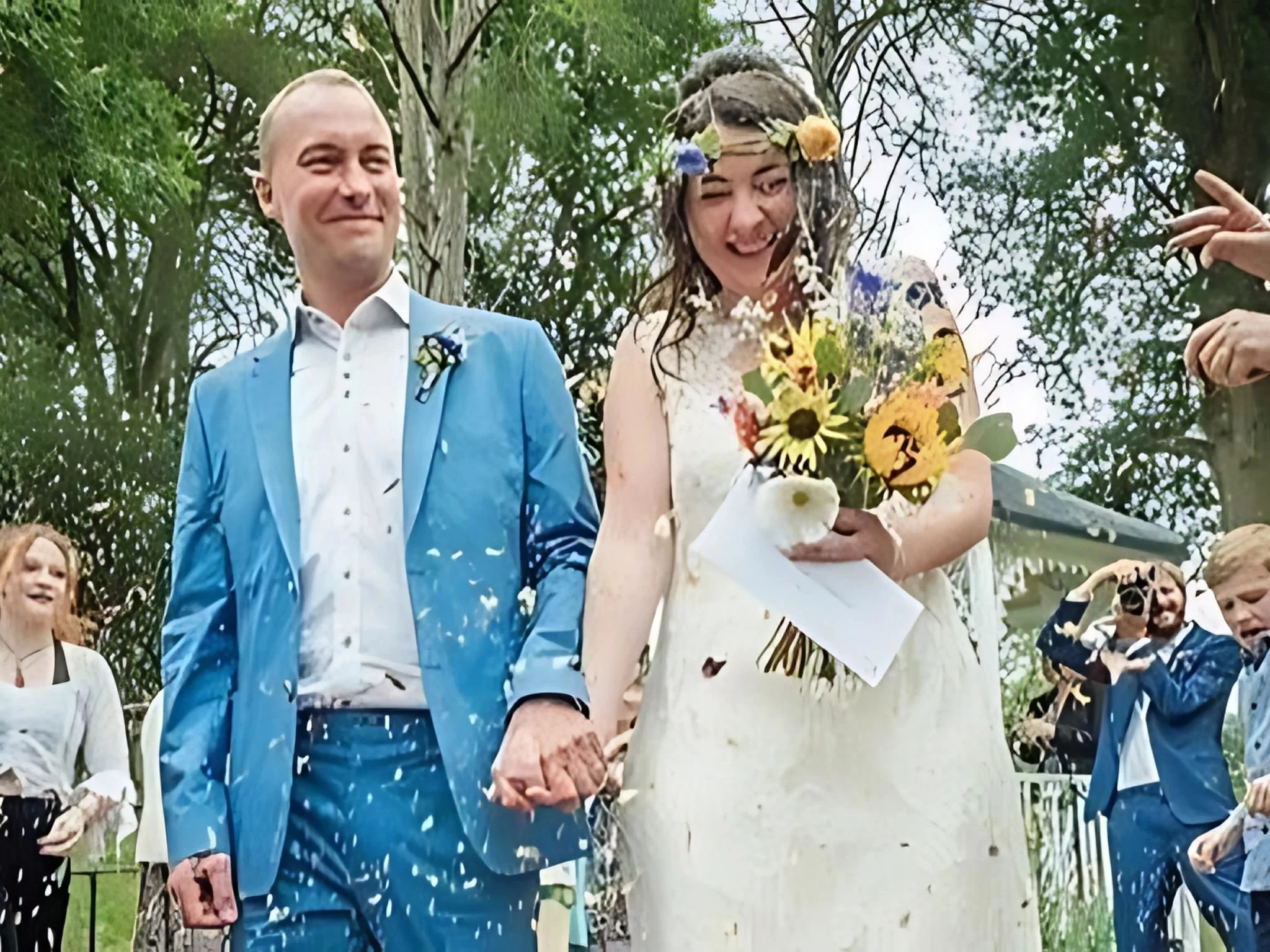 A bride and groom smiling and holding hands while walking outside. The groom is wearing a blue suit and the bride is in a white dress, holding a bouquet of flowers. People are celebrating and throwing something in the air around them.
