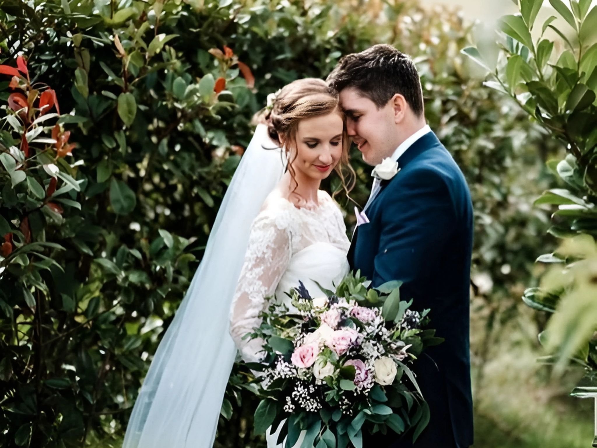 Bride and groom embracing in garden, bride holding floral bouquet, wearing white lace wedding dress and veil, surrounded by green foliage.