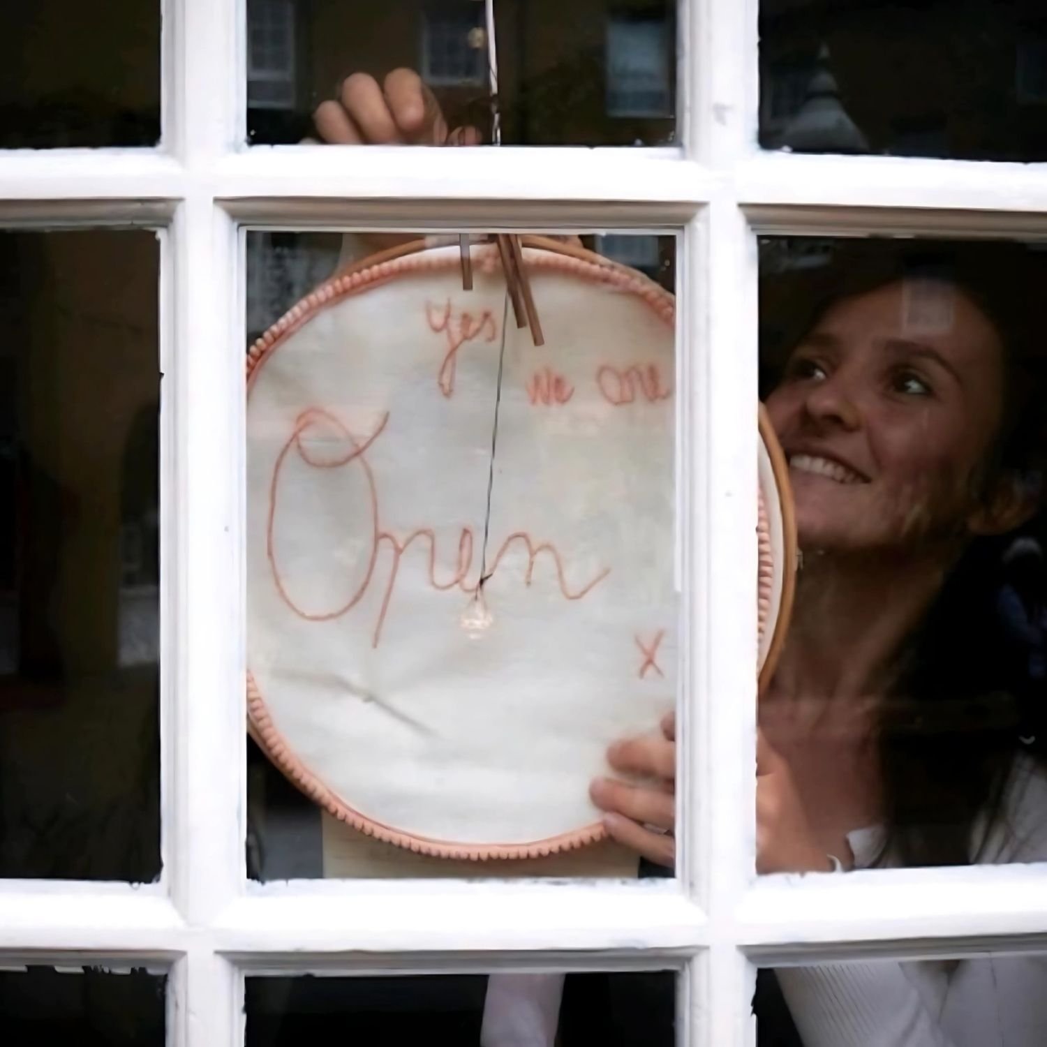 Person holding a hoop with an embroidered "Yes, we are Open" sign in a window.