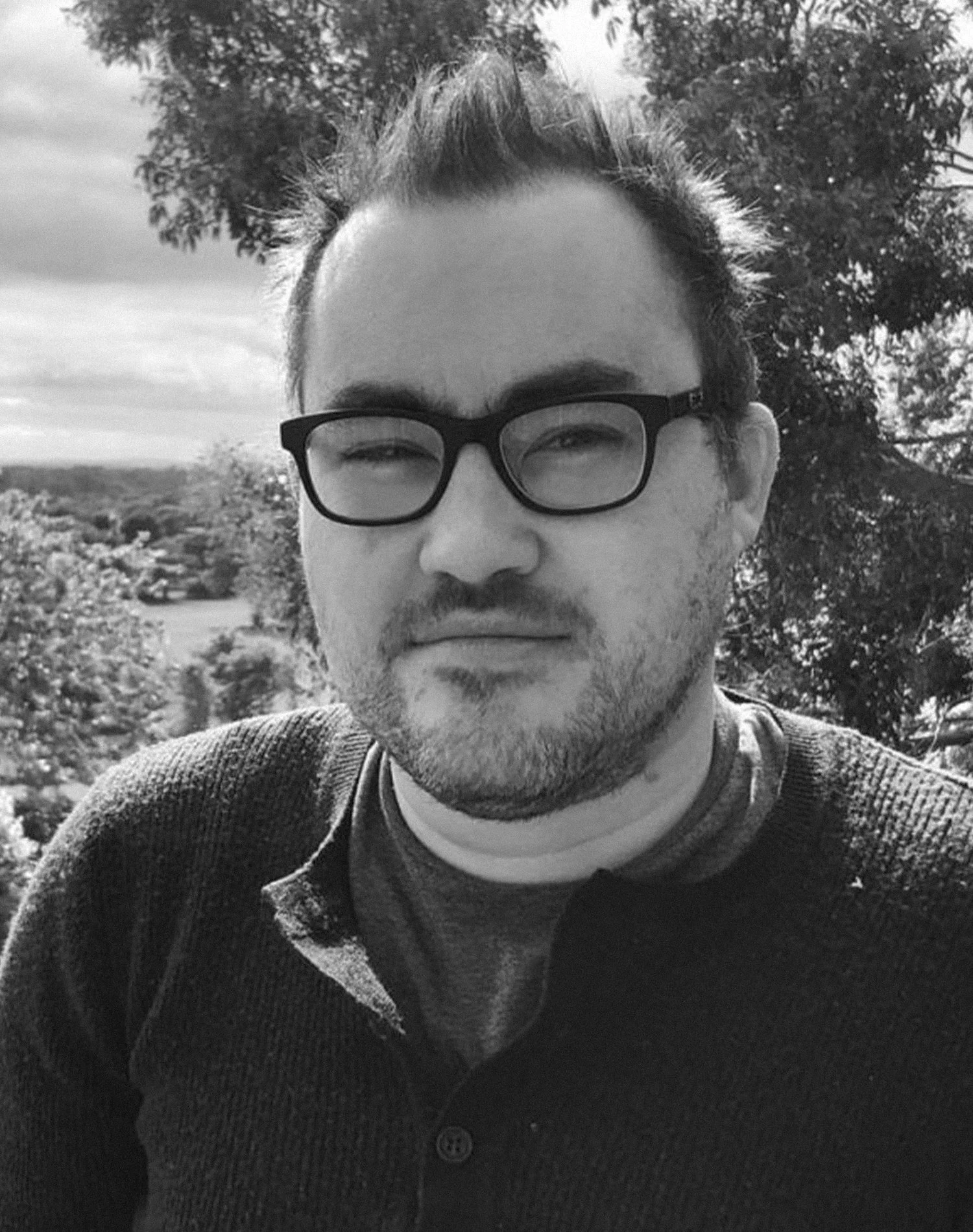 Black and white photo of a person wearing glasses outdoors in a natural setting with trees and a cloudy sky in the background.