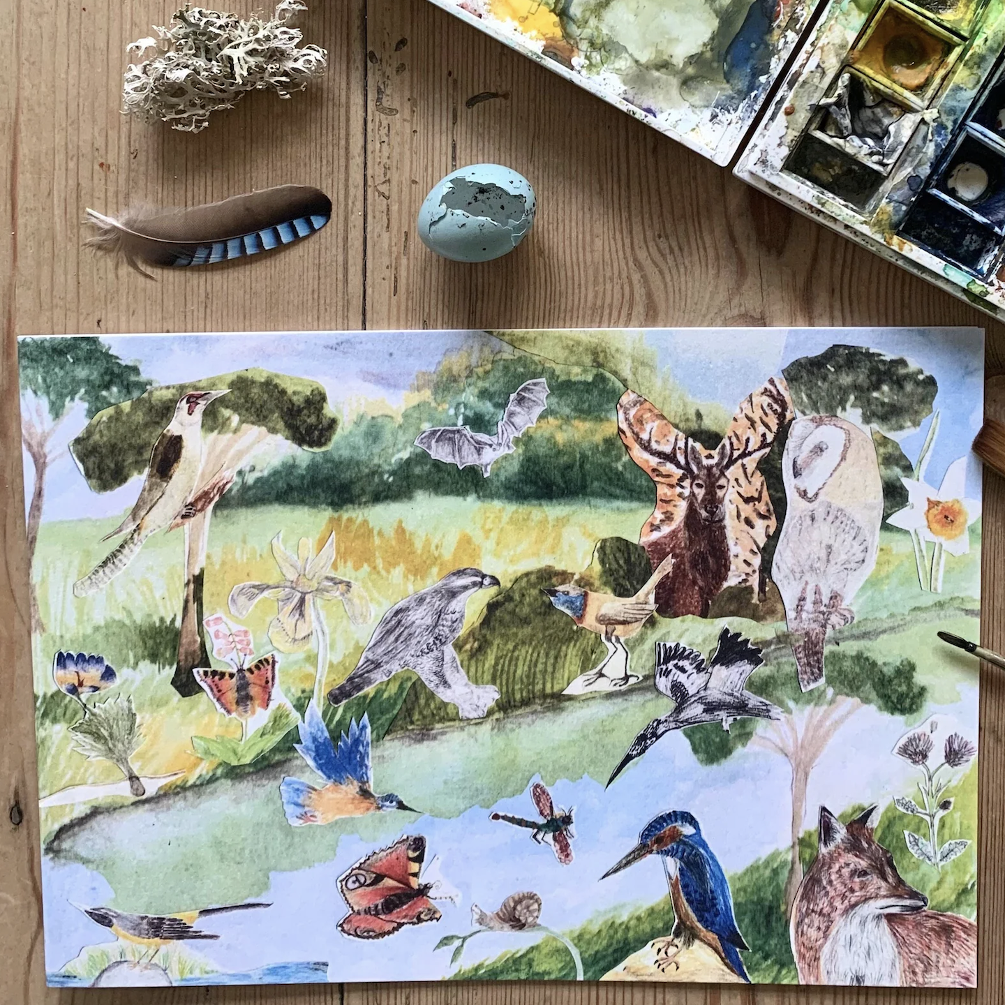 A watercolor painting depicting various wildlife in a natural setting, including birds, a bat, butterflies, and mammals. The image is surrounded by a paint palette, a blue cracked egg, a feather, and some natural elements like lichen on a wooden table.