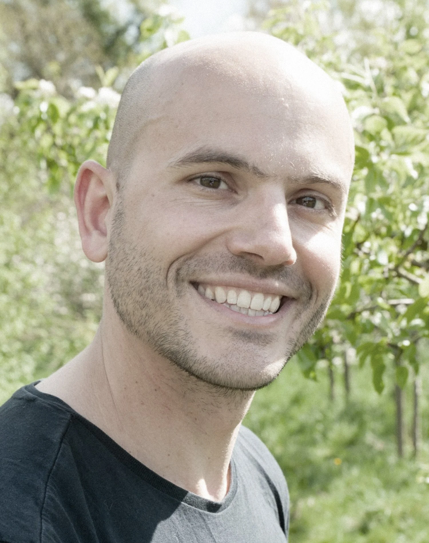 Bald man smiling outdoors with greenery in the background.