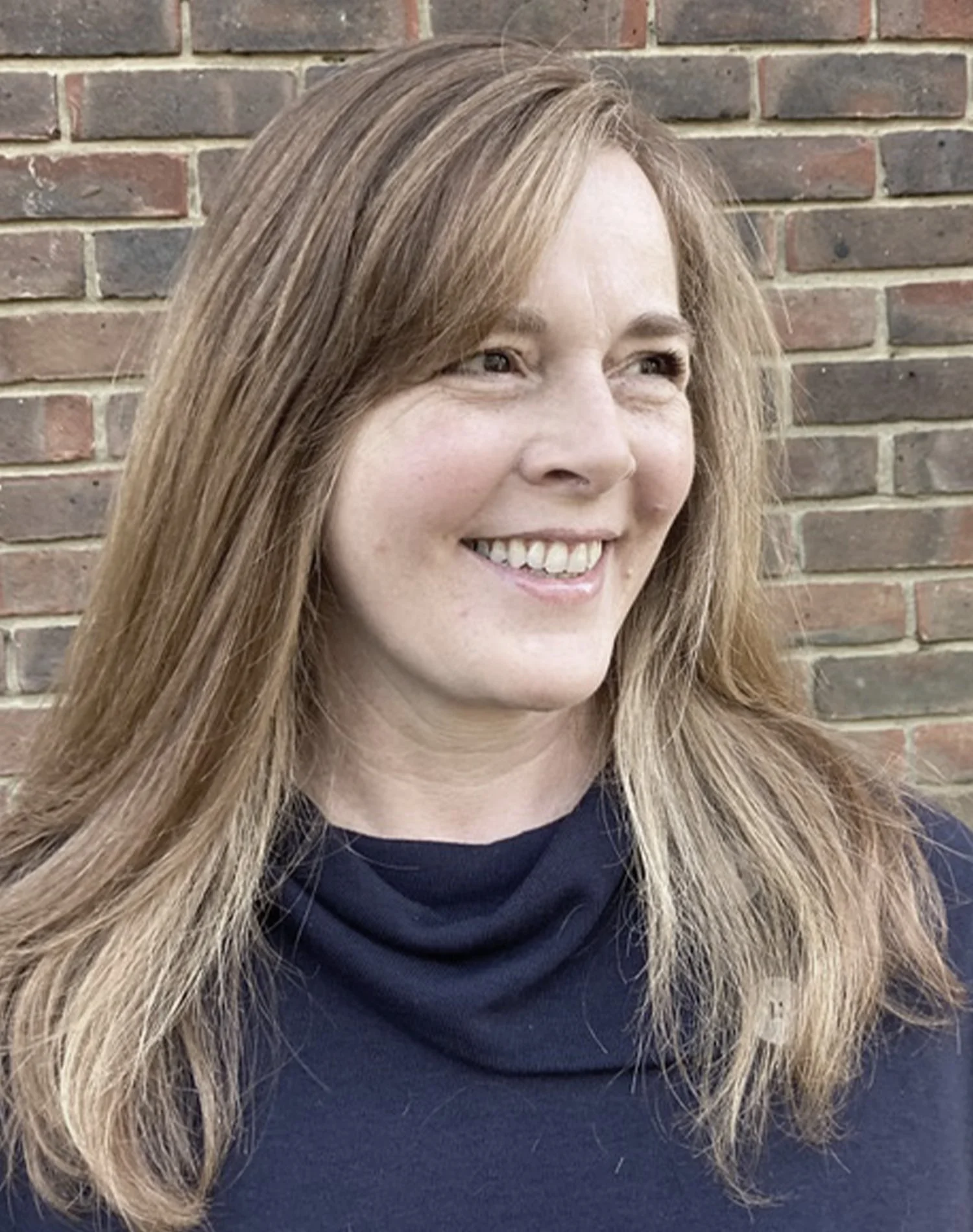 Woman with long hair smiling, standing against a brick wall.