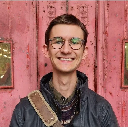Person with glasses smiling in front of a pink, weathered door.