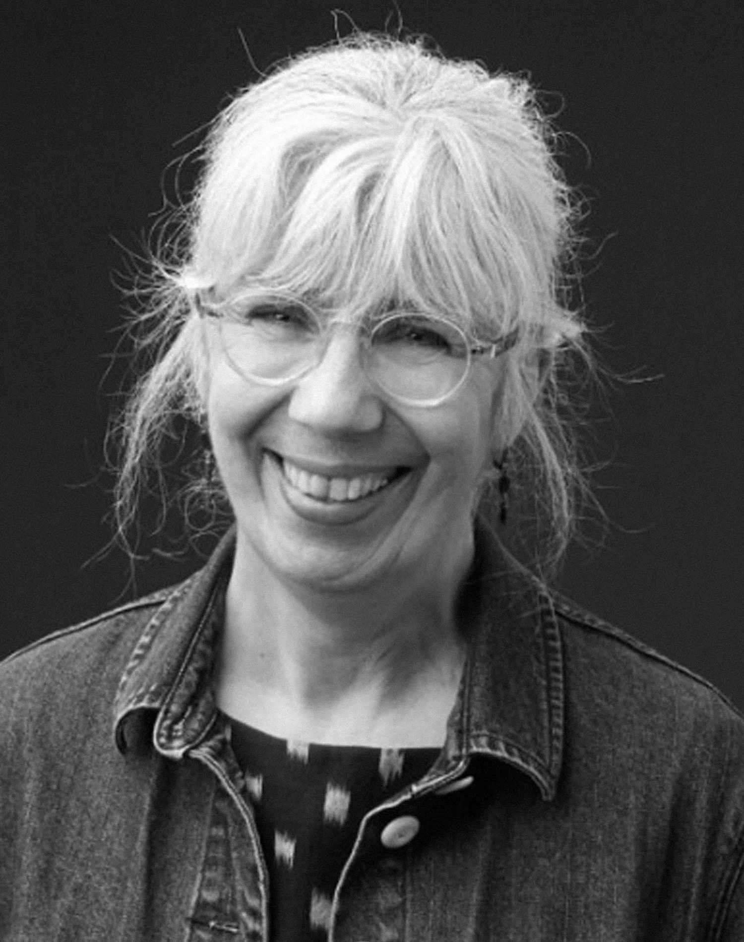 Black and white portrait of a smiling person wearing glasses and a denim jacket.