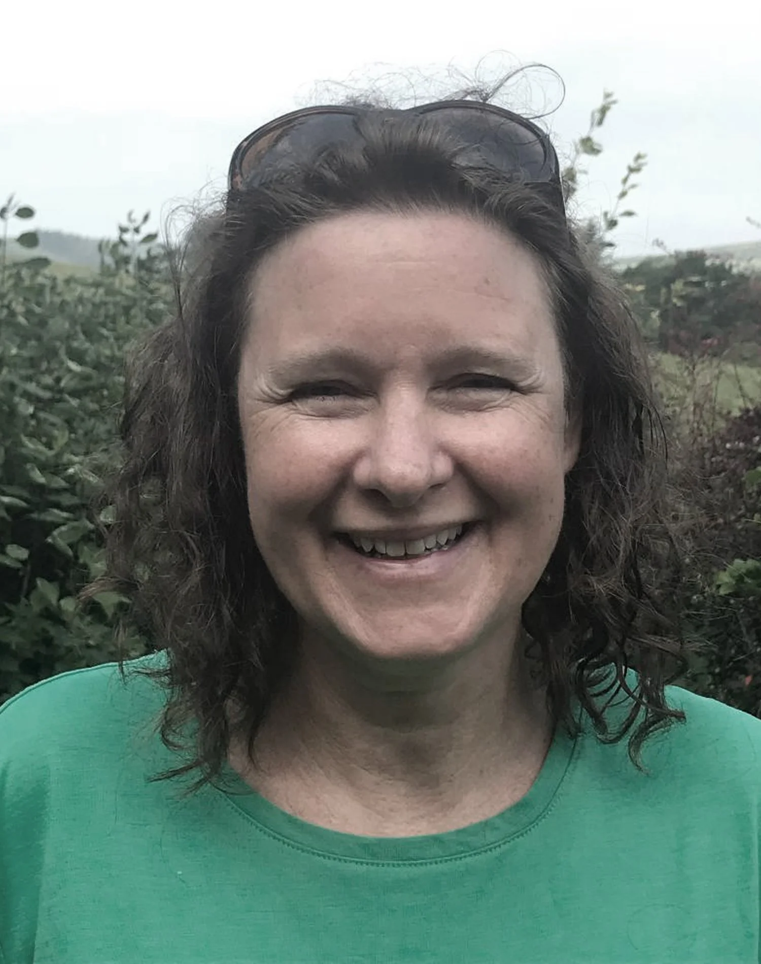 A smiling woman wearing a green shirt and sunglasses on her head, standing outdoors with greenery in the background.