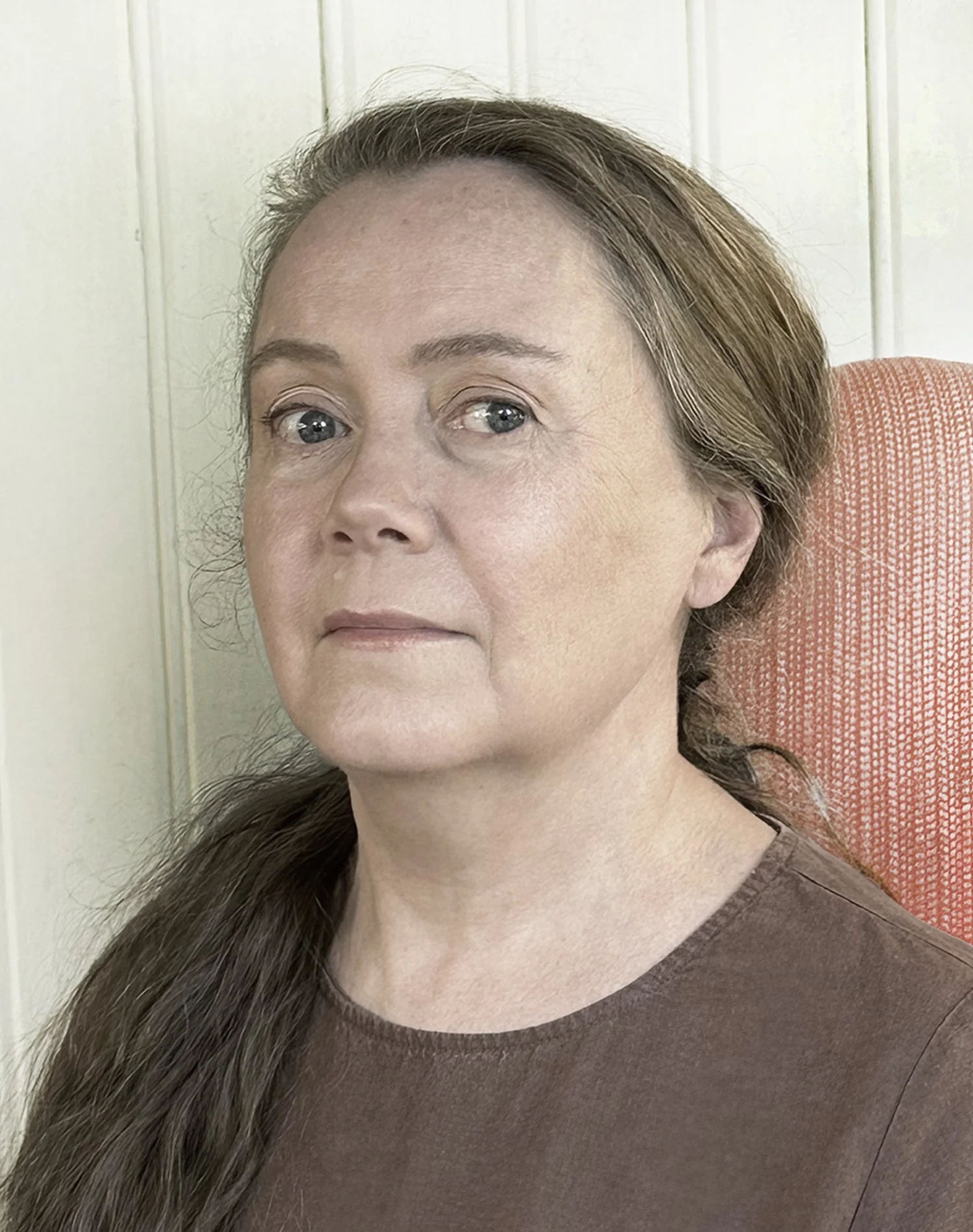 A person with long brown hair tied back, wearing a brown top, sitting on an orange chair against a light-colored wall.