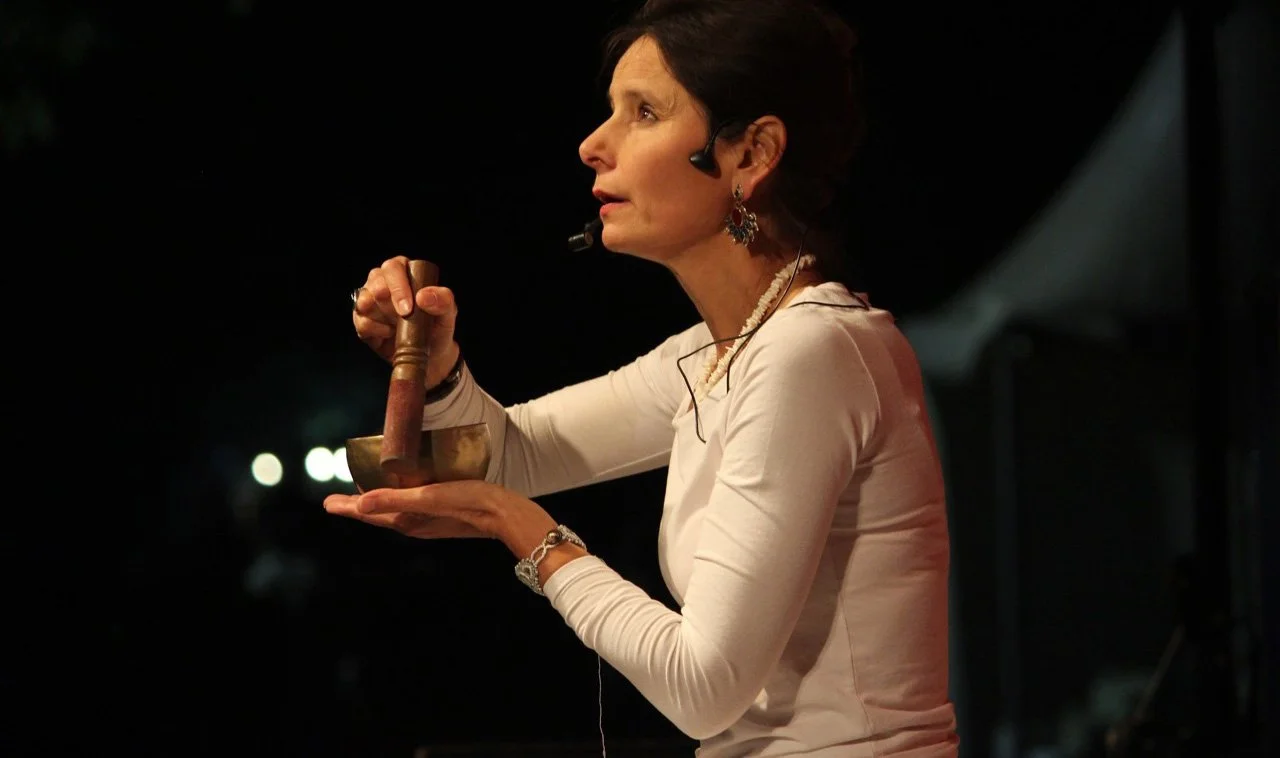 Person using a singing bowl with a mallet while wearing a headset microphone, against a dark background.