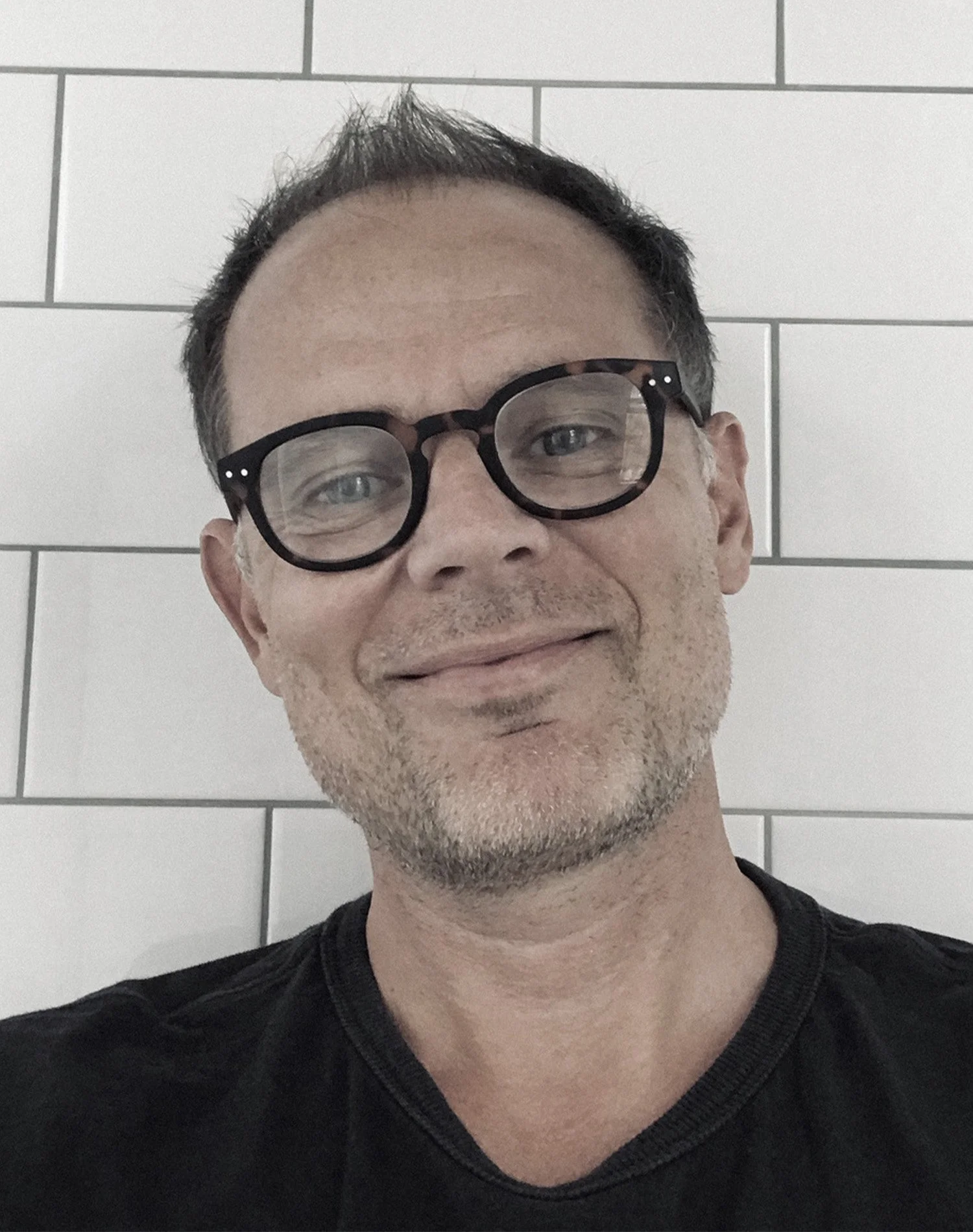 Person wearing black-framed glasses smiling in front of a white tiled wall.