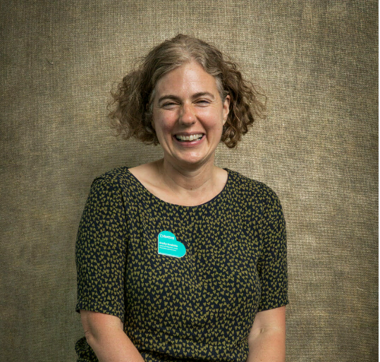 Smiling person with curly hair wearing a dark patterned shirt and a blue name tag against a burlap-textured background.