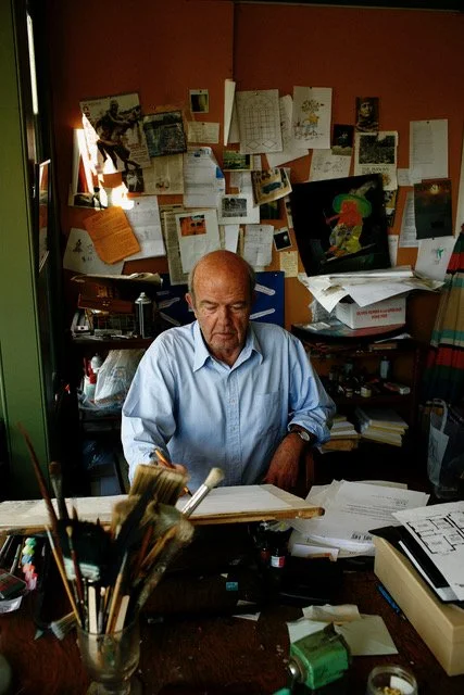 Man sitting at a cluttered desk with art supplies and papers, surrounded by a bulletin board filled with notes and images, possibly in an artist's studio.