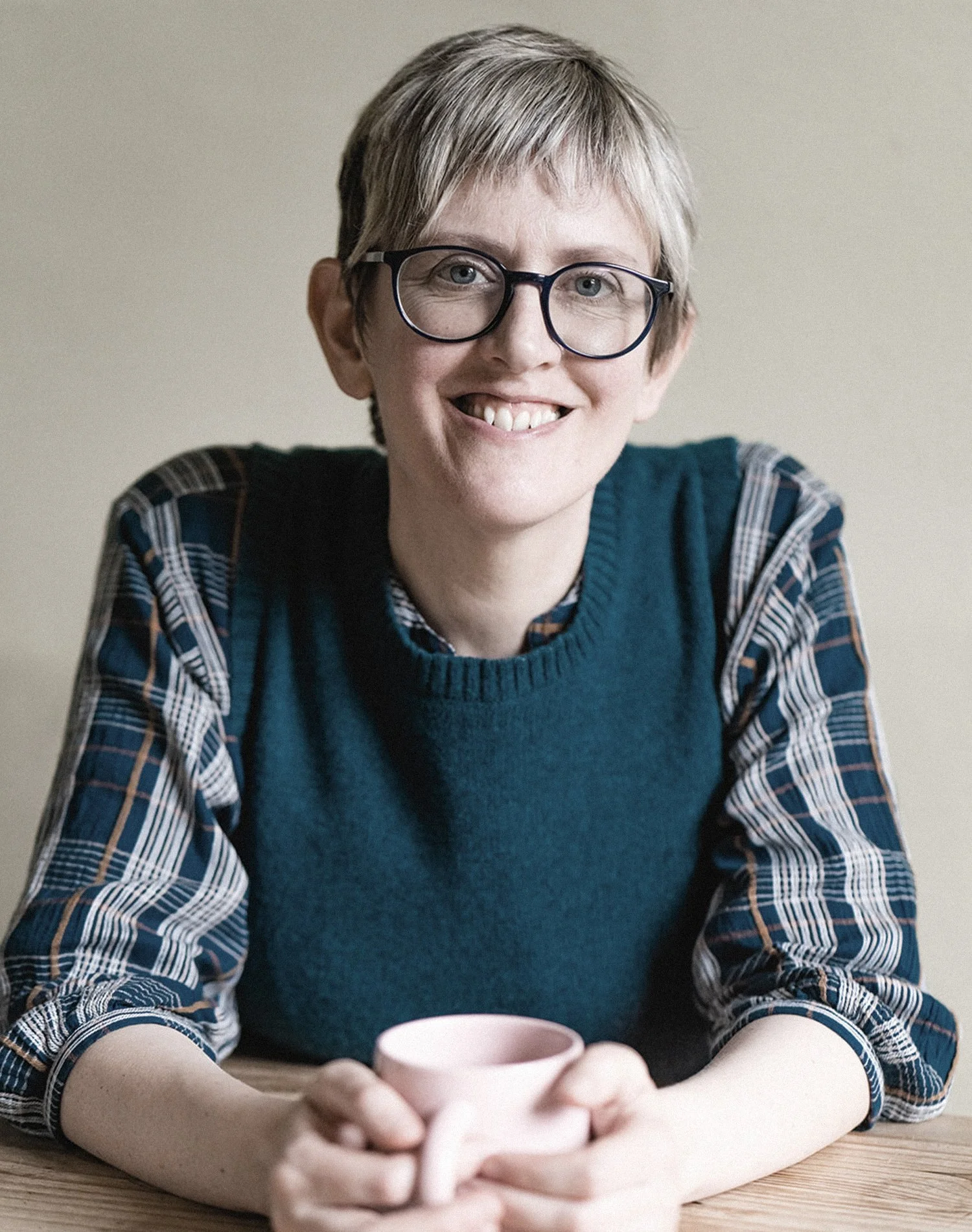 A person with short hair and glasses holding a pink mug, wearing a plaid shirt and a dark blue vest, smiling at the camera.