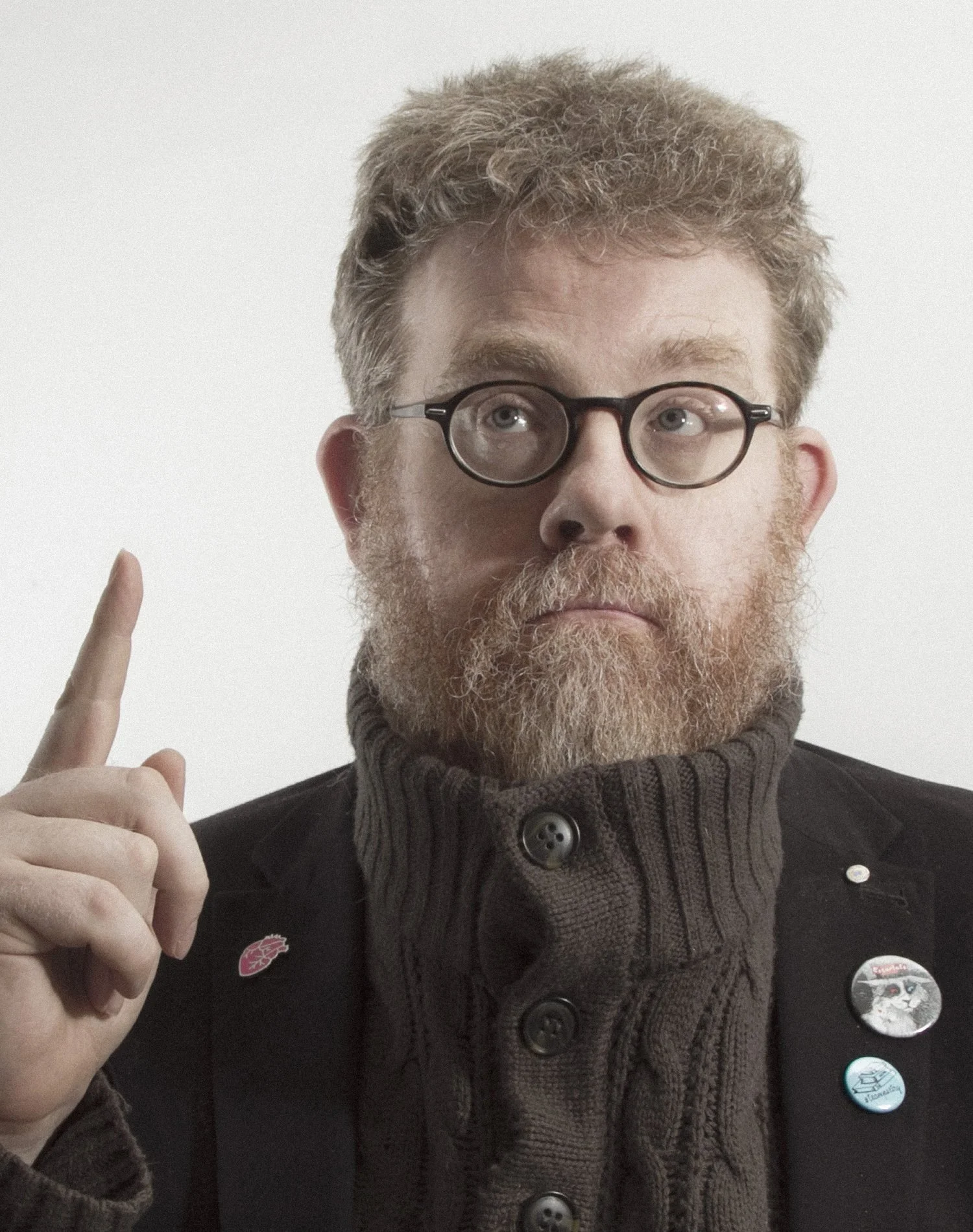 A person with glasses and a beard wearing a thick, buttoned sweater and a black jacket adorned with lapel pins, holding up one finger.