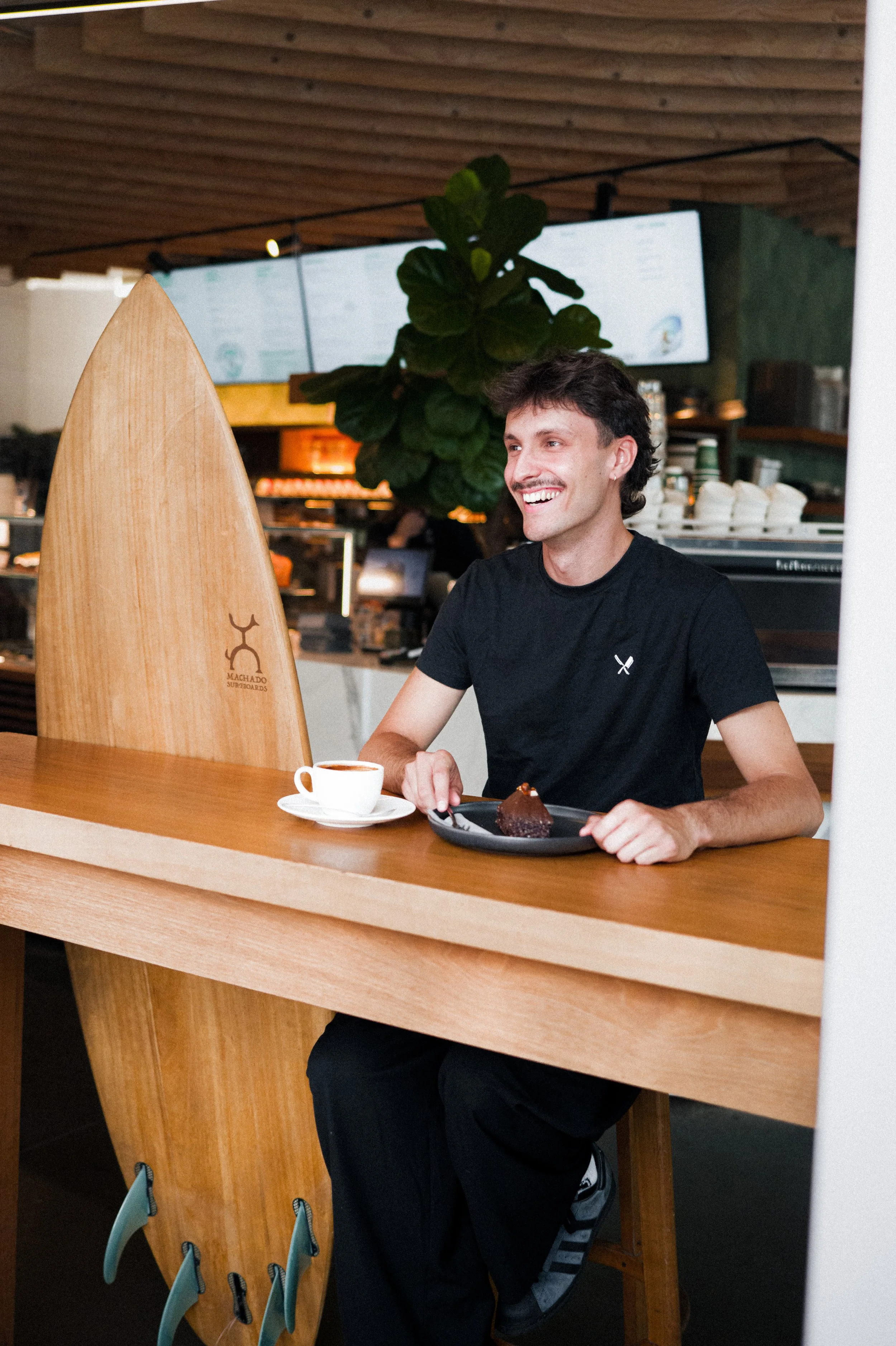 Ein lachender junger Mann sitzt an einem Holztresen im Café, vor ihm eine Tasse Kaffee und ein Stück Schokoladenkuchen auf einem Teller. Im Hintergrund befindet sich eine große grüne Pflanze und eine Menütafel.