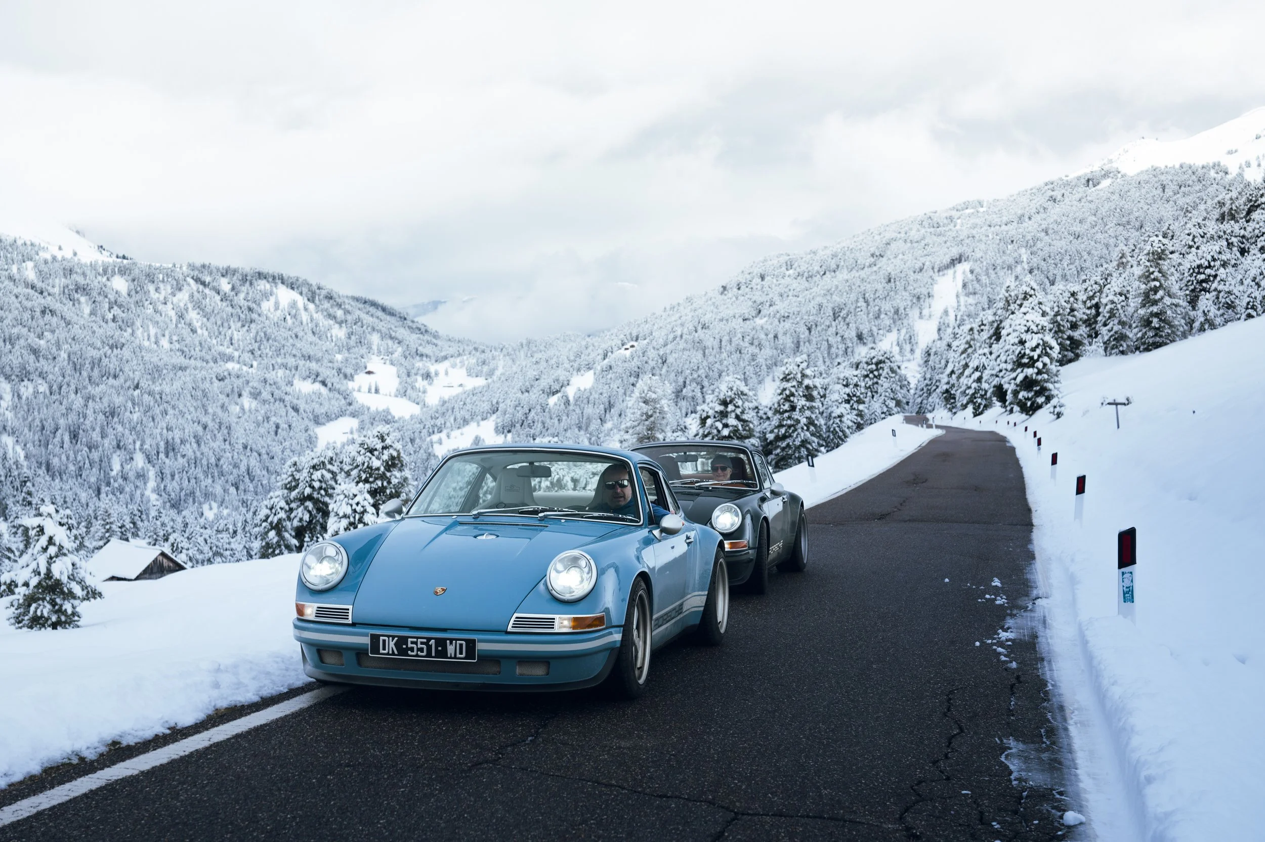 Zwei Porsche-Sportwagen, ein blauer und ein schwarzer, fahren auf einer schneebedeckten Bergstraße. Die Landschaft ist mit verschneiten Bäumen und Bergen im Hintergrund. Die Fahrer tragen Sonnenbrillen, und der Himmel ist bewölkt.