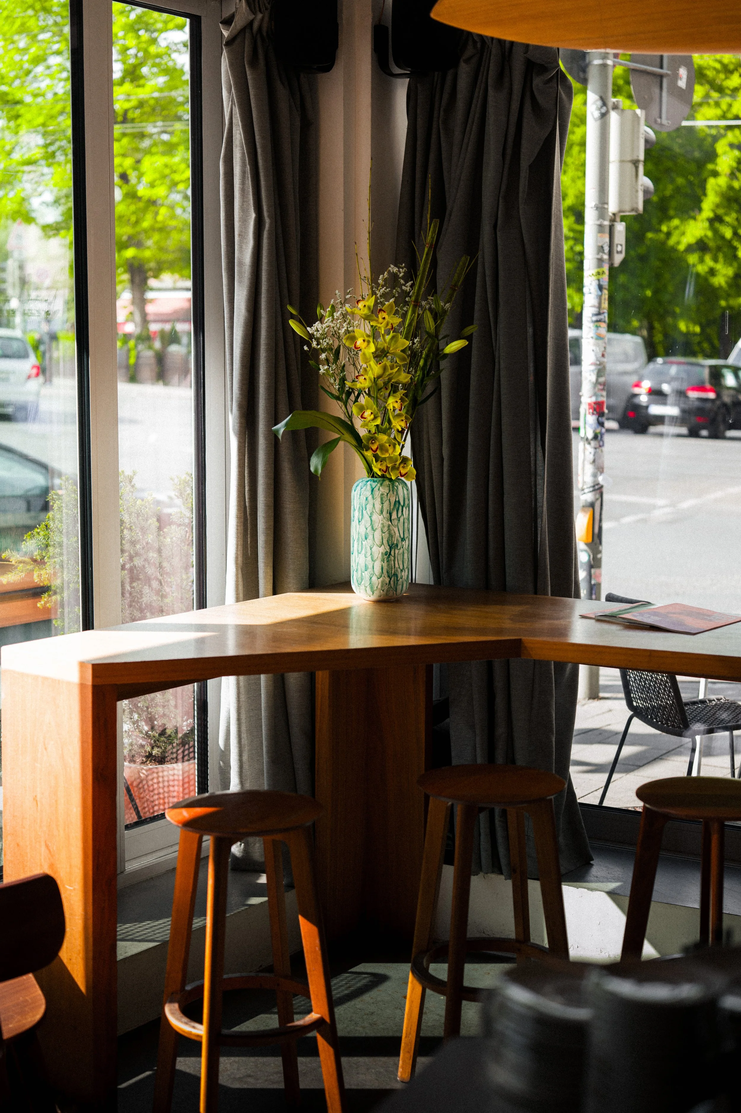 Ein Tisch mit drei Stühlen, einer Vase mit gelben Orchideen und weißen Blumen vor einem großen Fenster mit grauen Vorhängen. Draußen sind Bäume, Autos und eine Straßenlaterne sichtbar.