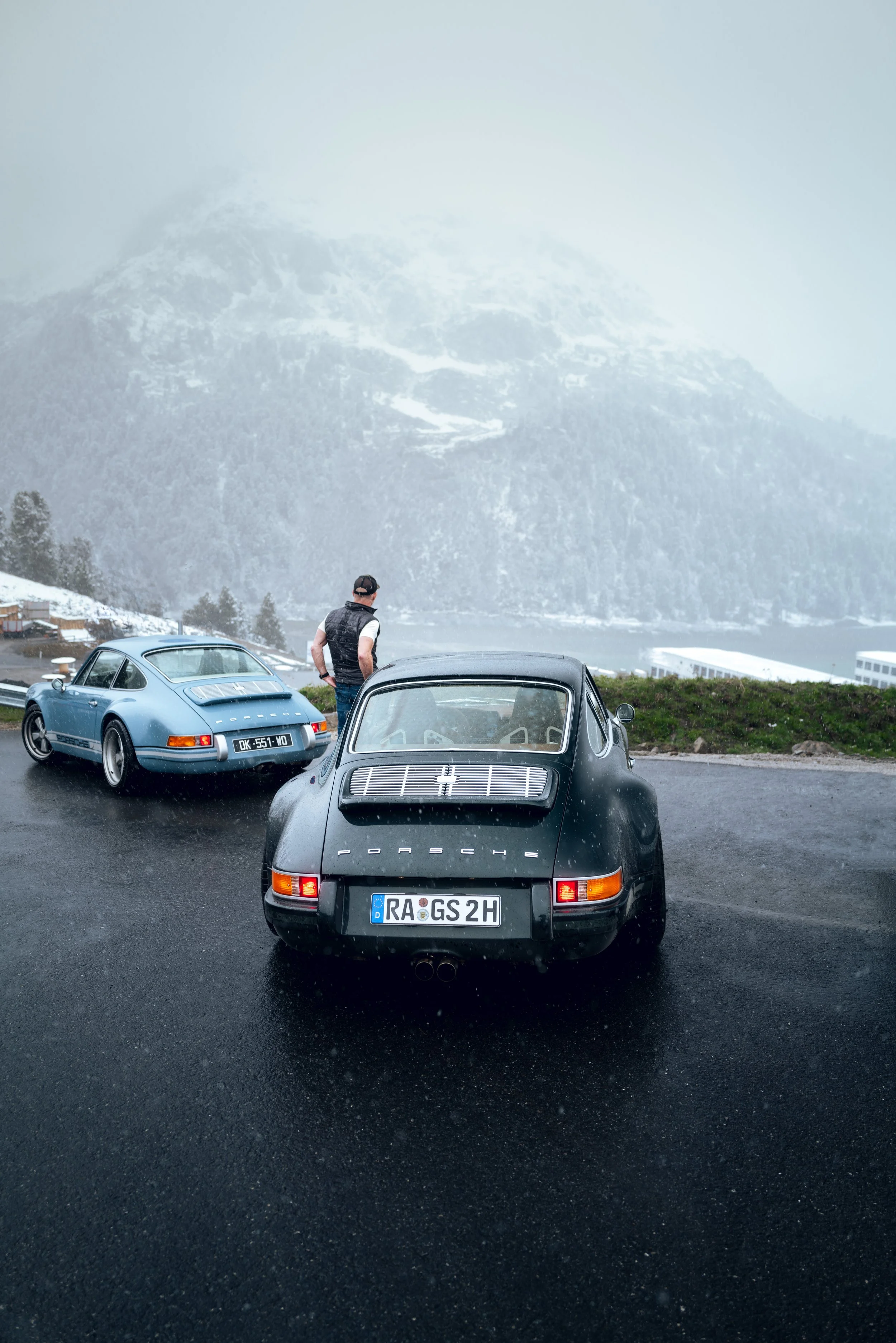 Zwei Porsche-Sportwagen, ein schwarzer und ein blauer, sind auf einer regennassen Straße vor einer Bergkulisse mit Schneefall und Nebel geparkt, während ein Mann in einer Jacke zwischen den Fahrzeugen steht.
