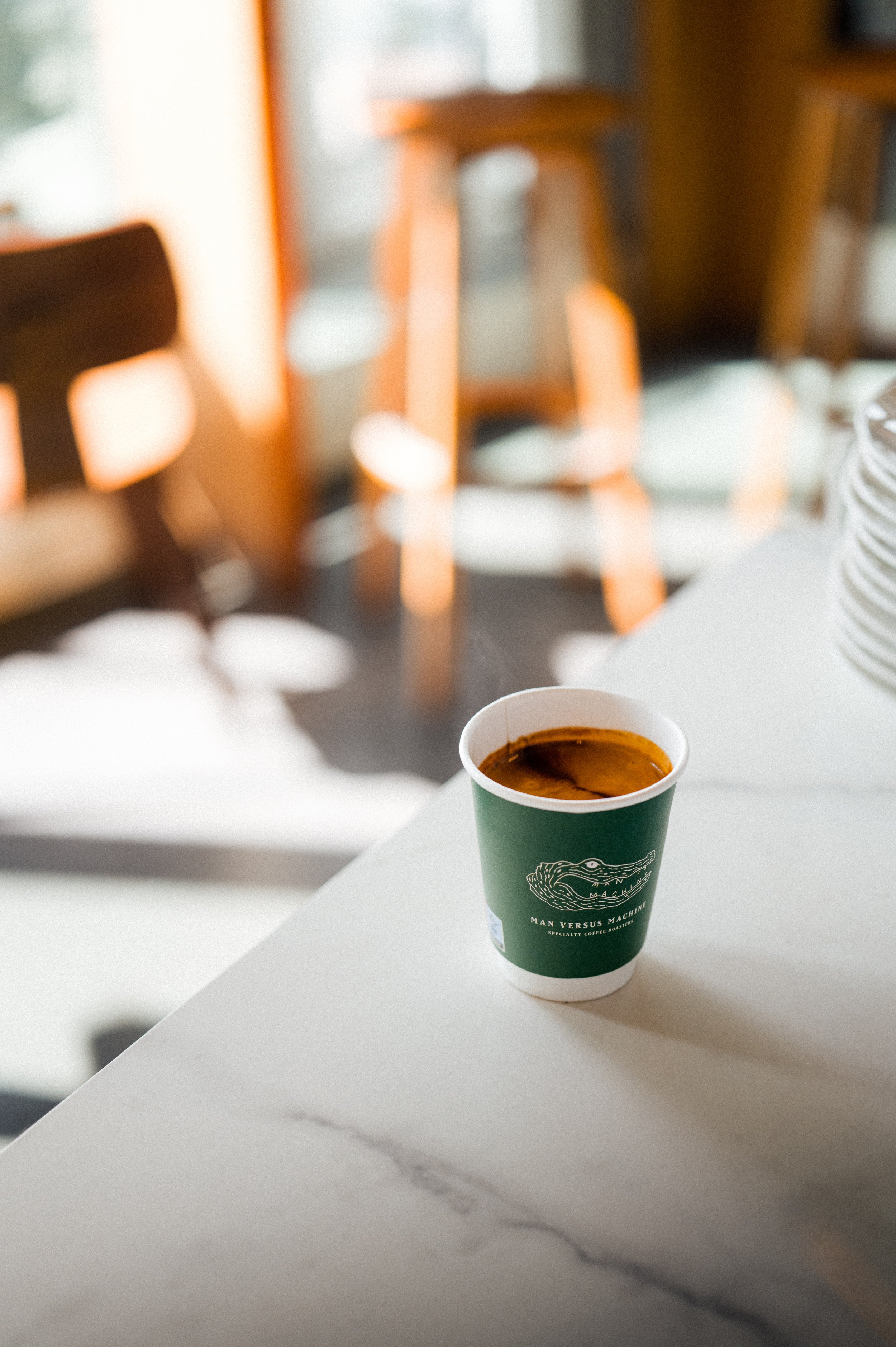 Ein Becher Kaffee auf einem weißen Tisch, im Hintergrund unscharfe Holzstühle und Möbel.