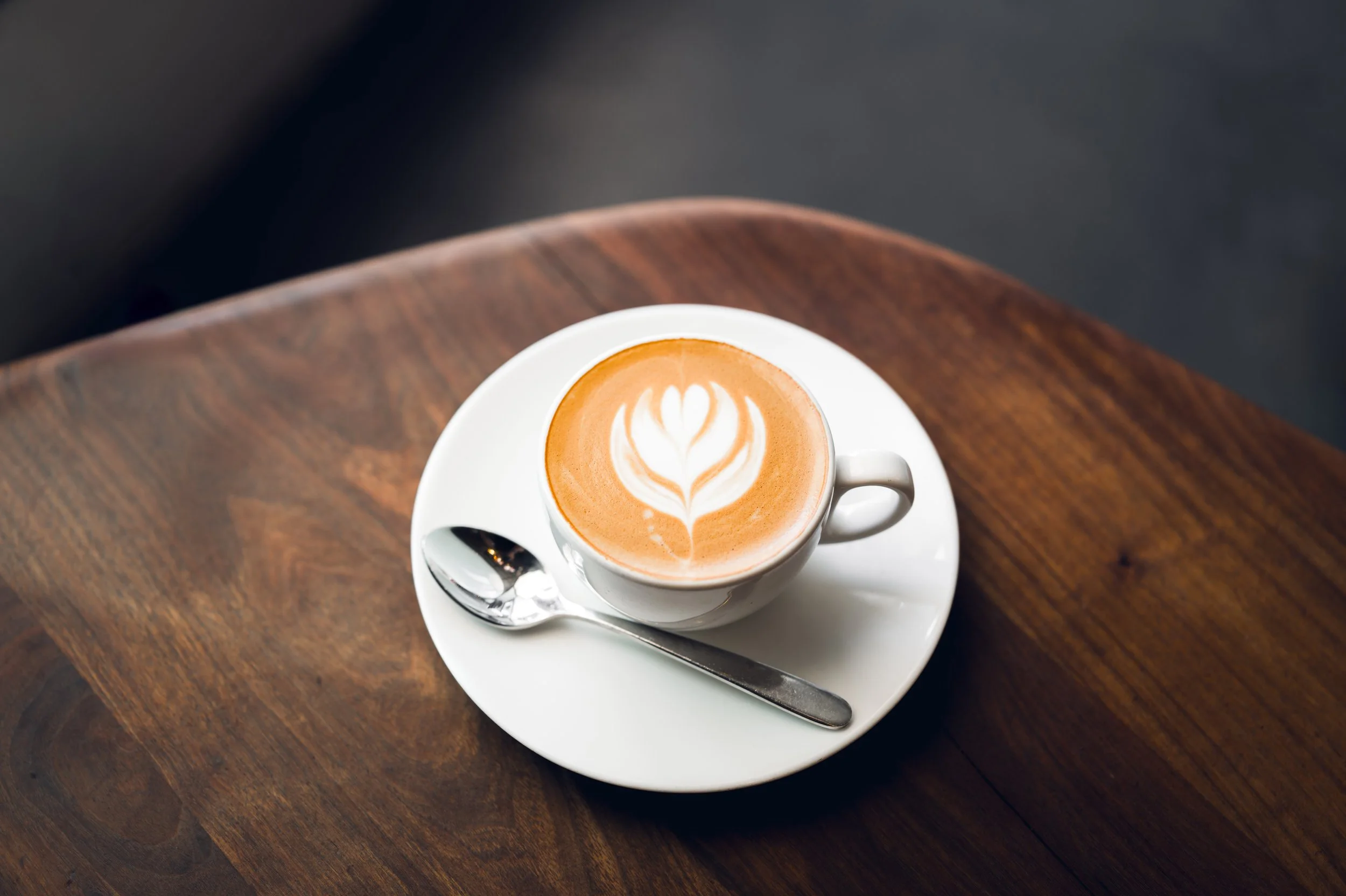 Eine Tasse Cappuccino mit Latte-Art auf einem weißen Unterteller, auf einem dunklen Holztisch, mit einem Löffel daneben.