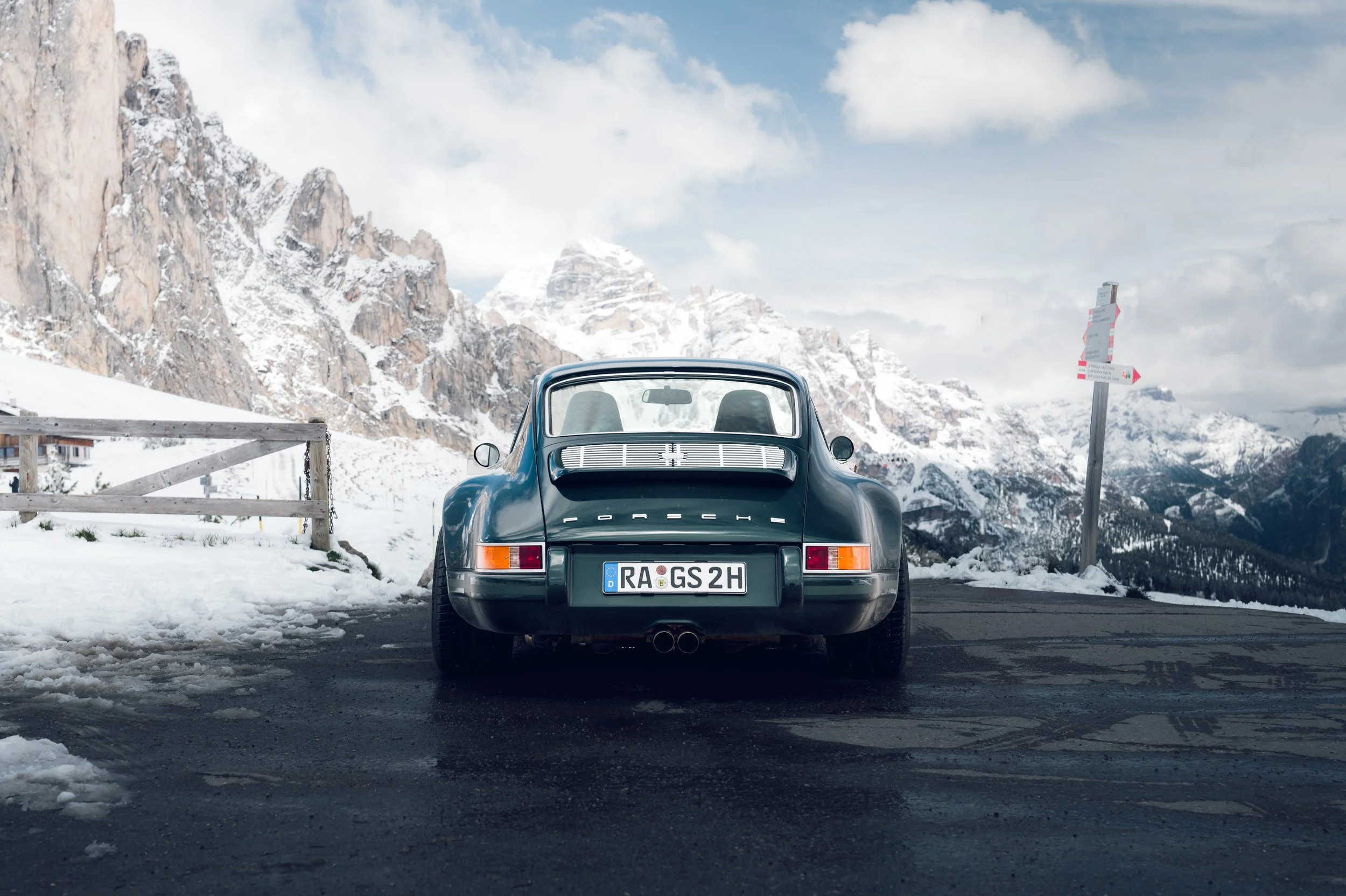 Ein schwarzer Porsche-Sportwagen steht auf einer schneebedeckten Straße in den Bergen. Im Hintergrund sind hohe, schneebedeckte Berge und ein Teil des blauen Himmels mit Wolken sichtbar.