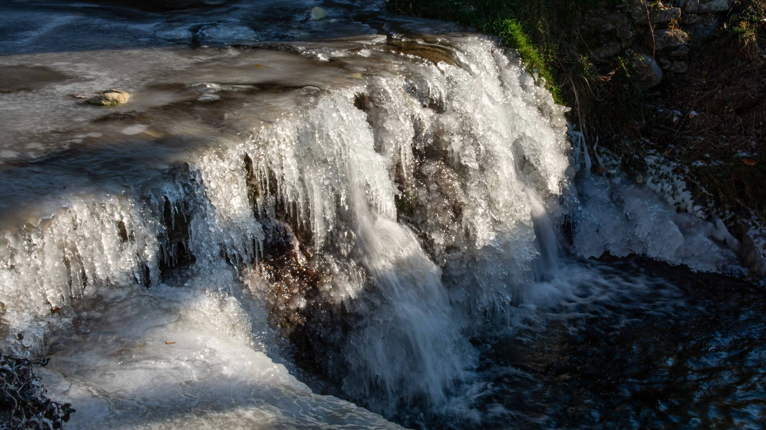 Une cascade gelée avec de la glace et de l'eau qui coule, entourée de végétation et de pierres.