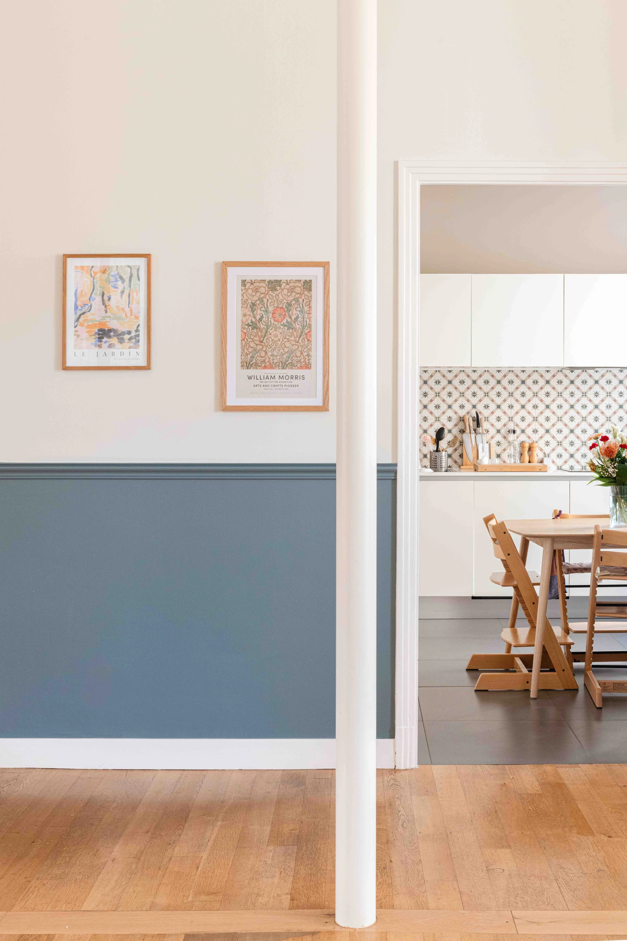 Vue de l'intérieur d'une maison avec une séparation architecturale entre une partie salon avec murs gris clair et une cuisine claire avec carrelage à motif, tables en bois et chaises.