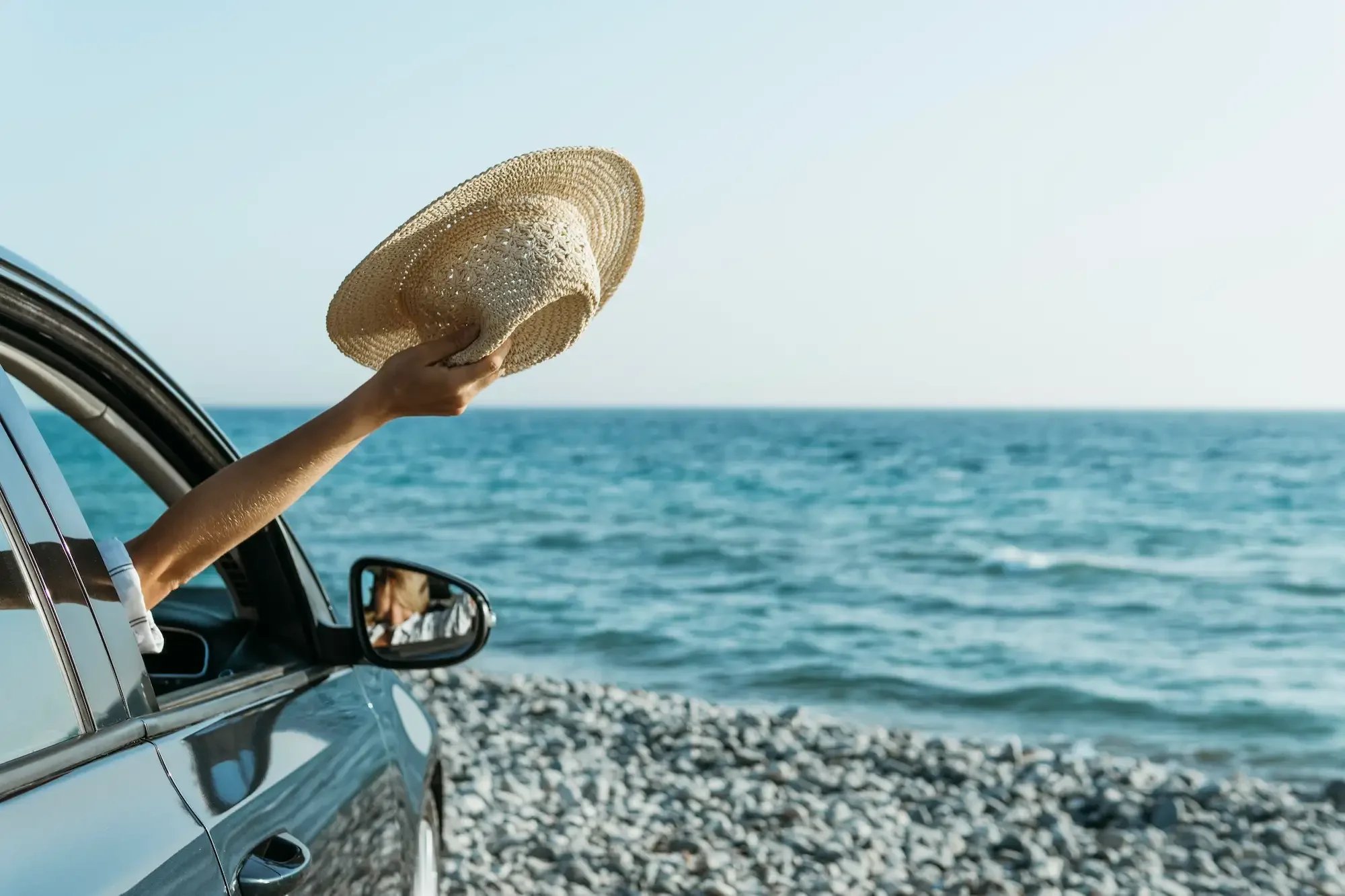 A woman holding a straw hat out of a rental car window while overlooking the Mediterranean coast in Cyprus.