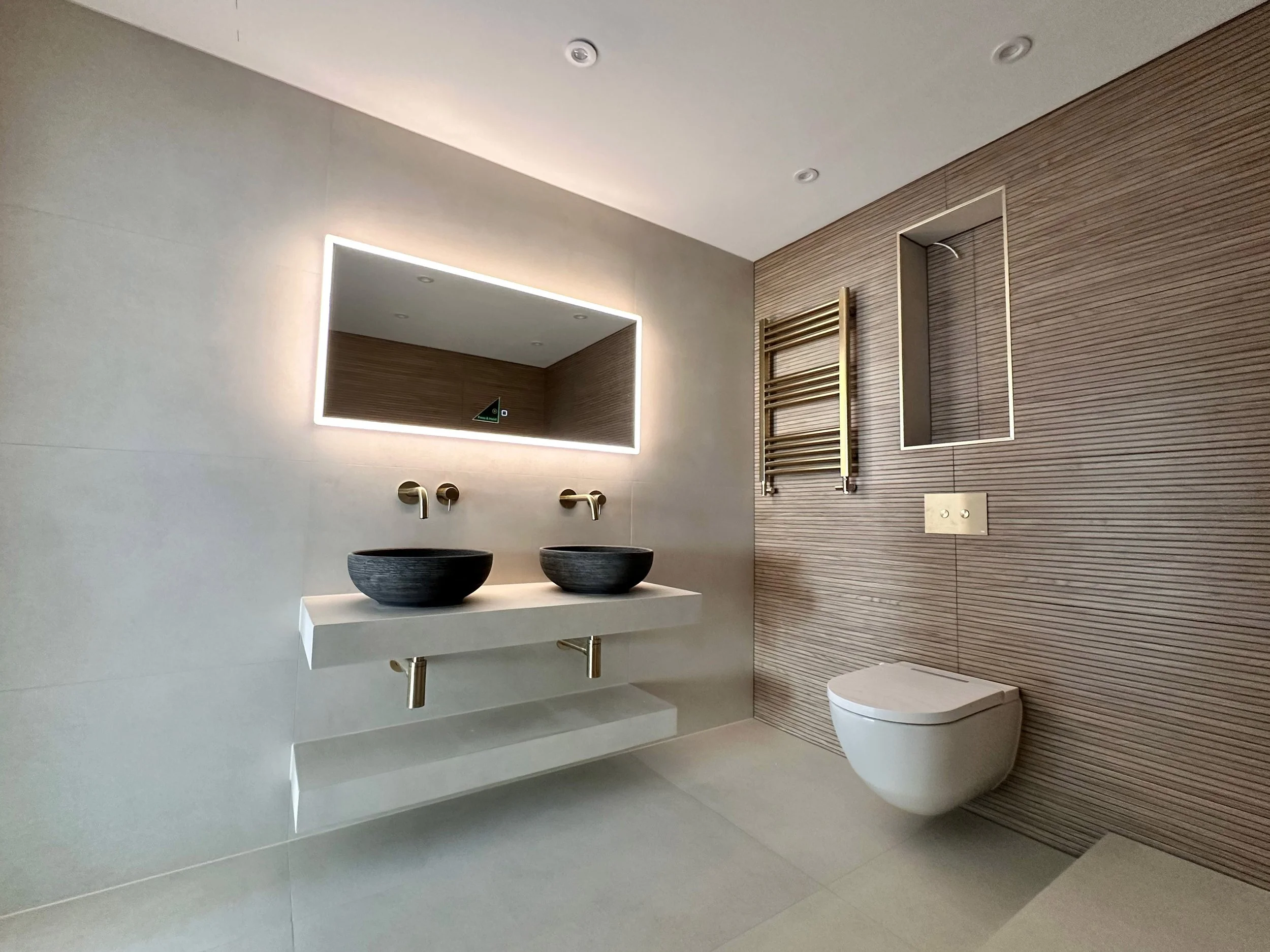 Modern bathroom with dual black vessel sinks, a large backlit mirror, a wall-mounted toilet, a heated towel rail, and a textured wood accent wall.