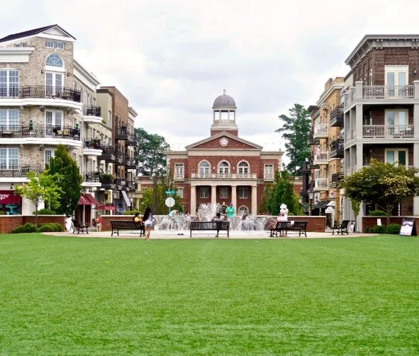 A park with green grass, benches, and fountains in front of a red brick building with a clock tower, surrounded by residential buildings.