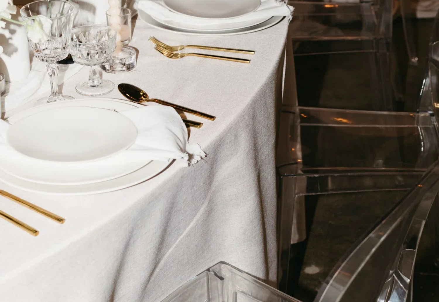 Elegant table setting with white plates, gold cutlery, crystal glasses, and a white tablecloth, beside transparent chairs.