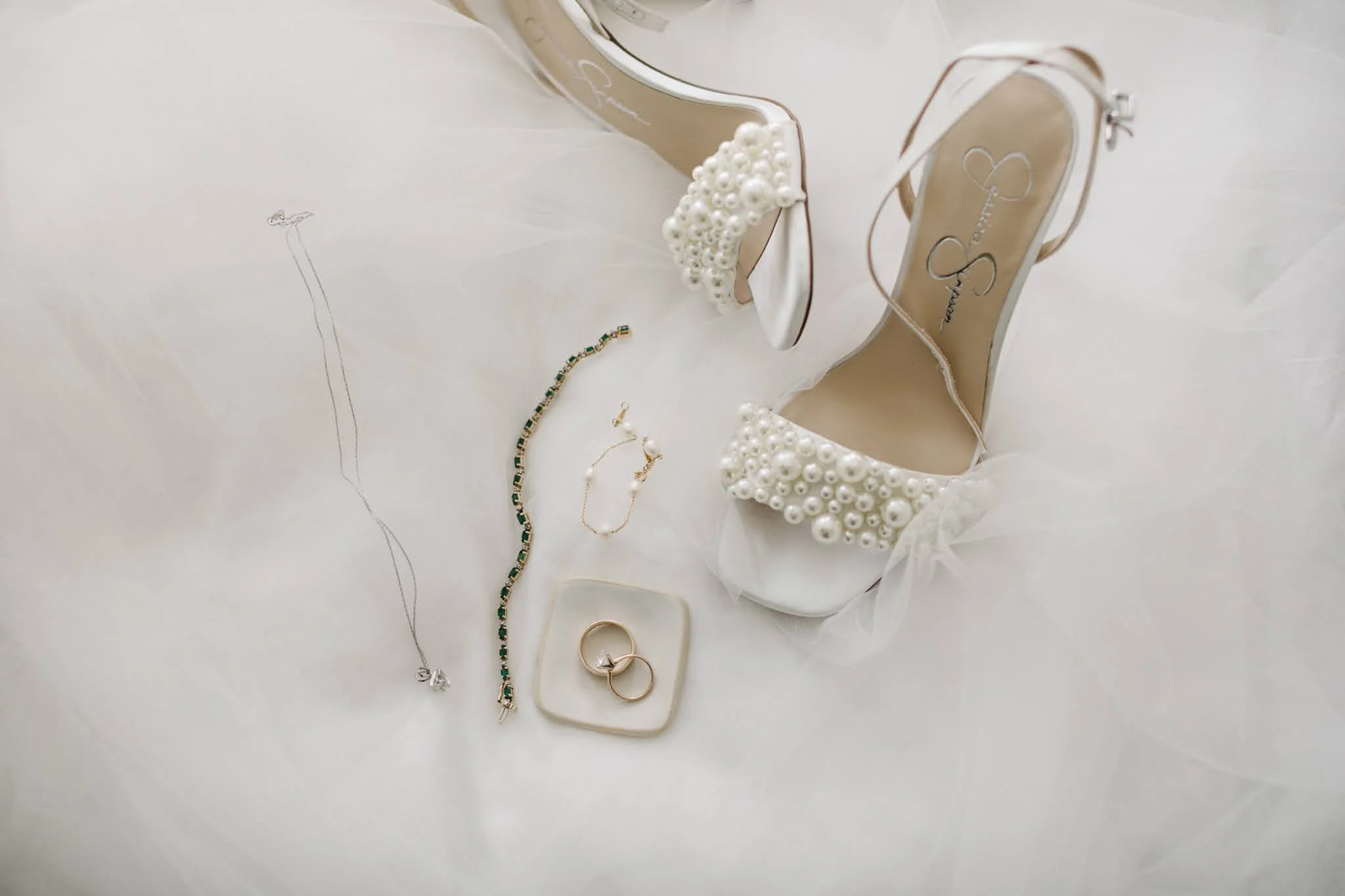 A pair of white bridal shoes with pearl embellishments, a pearl necklace, a pair of pearl and gold earrings, a gold ring with two rings on a white dish, and a gold bracelet with green accents, all placed on a white fabric background.